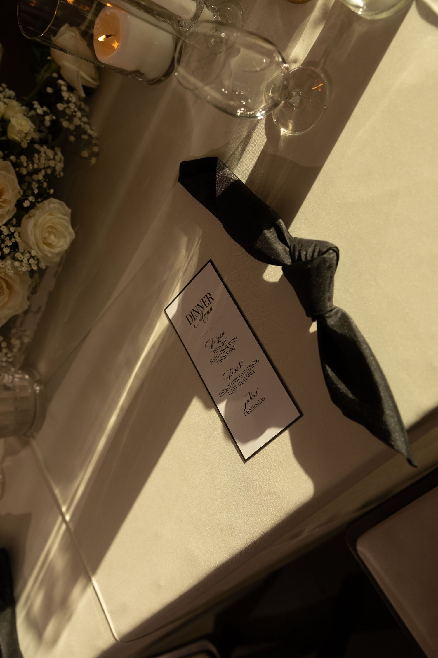 Elegant wedding dinner menu card tied with black ribbon on white tablecloth with roses and candles.