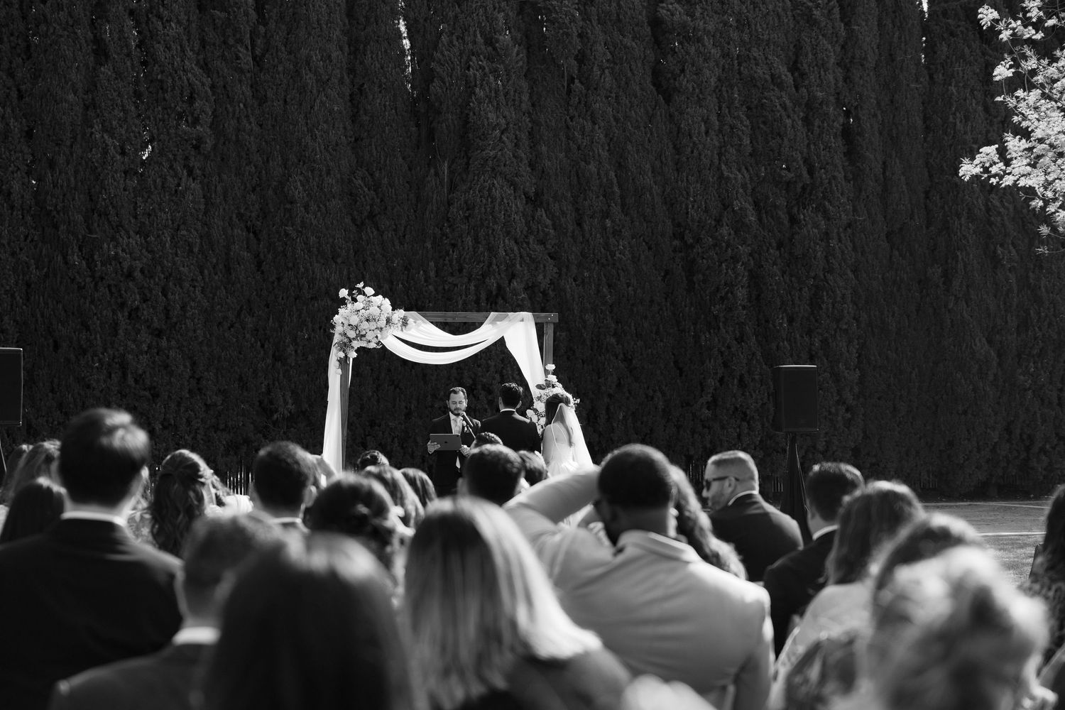 Guests watch as a couple exchanges vows at an outdoor wedding ceremony beneath a floral arch.