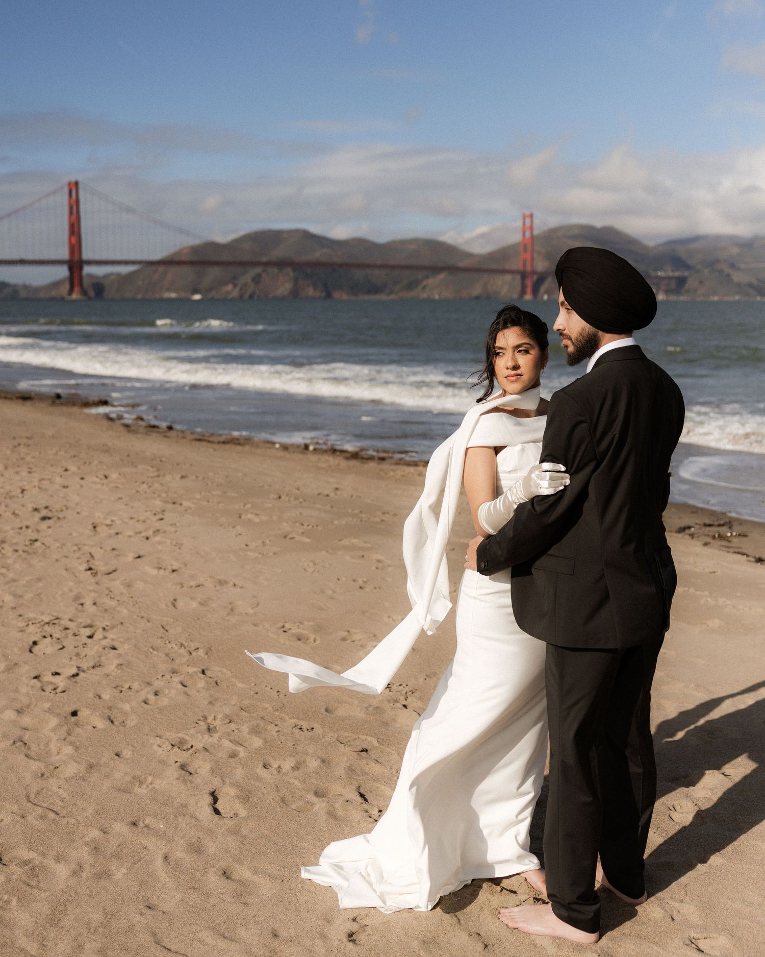 Two figures in contrasting white and black outfits dance playfully on a beach with the Golden Gate Bridge in the background.