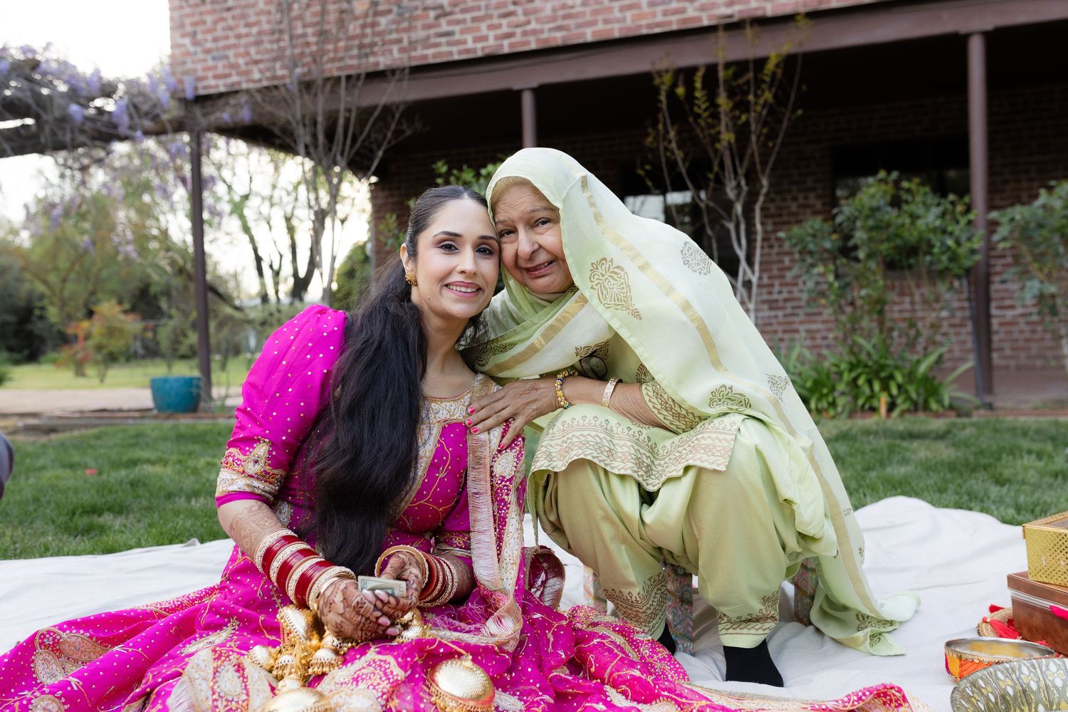 A couple in traditional South Asian wedding attire reclines on pink and gold decorative cushions outdoors.