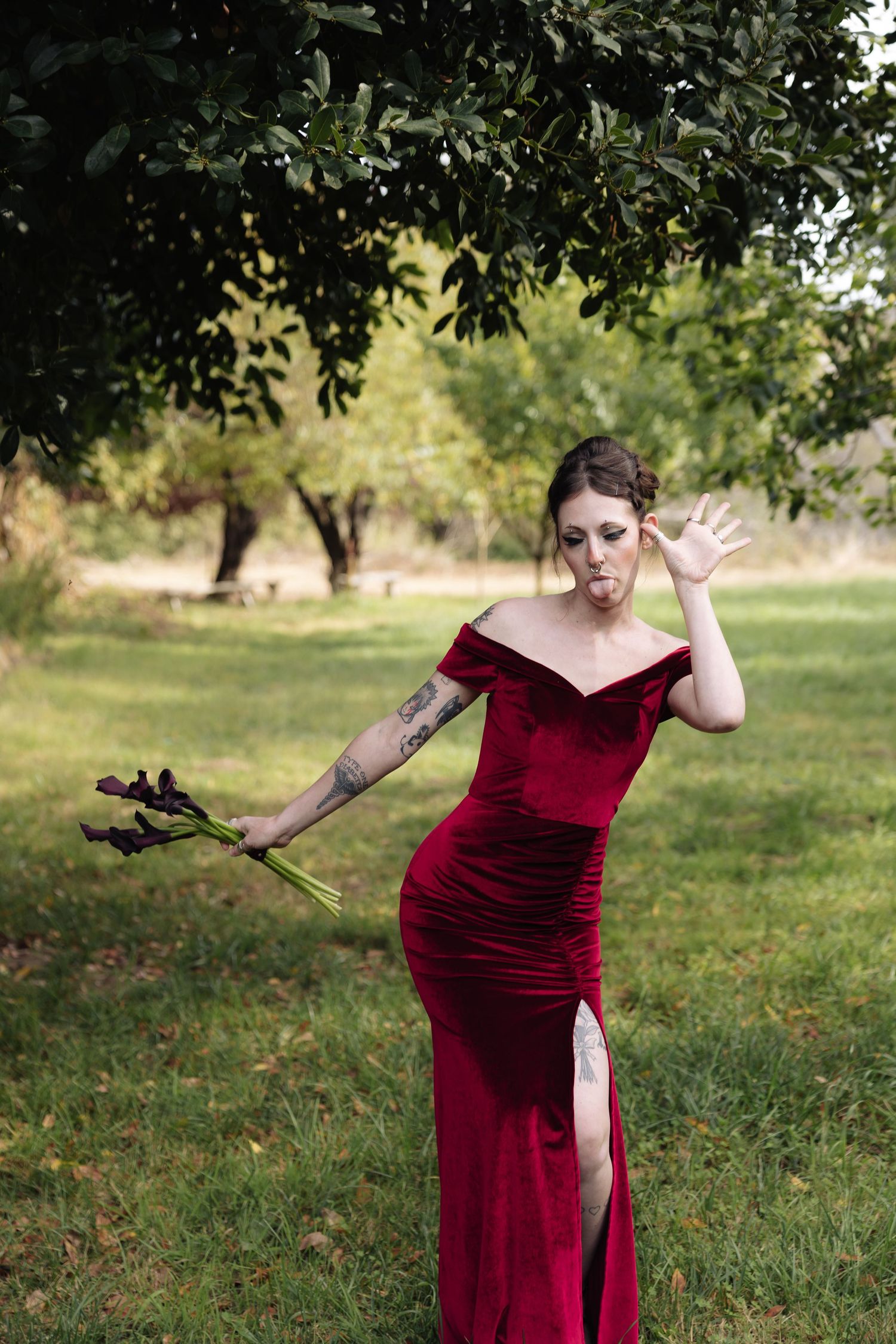 Figure in burgundy velvet gown poses dramatically against natural outdoor backdrop.