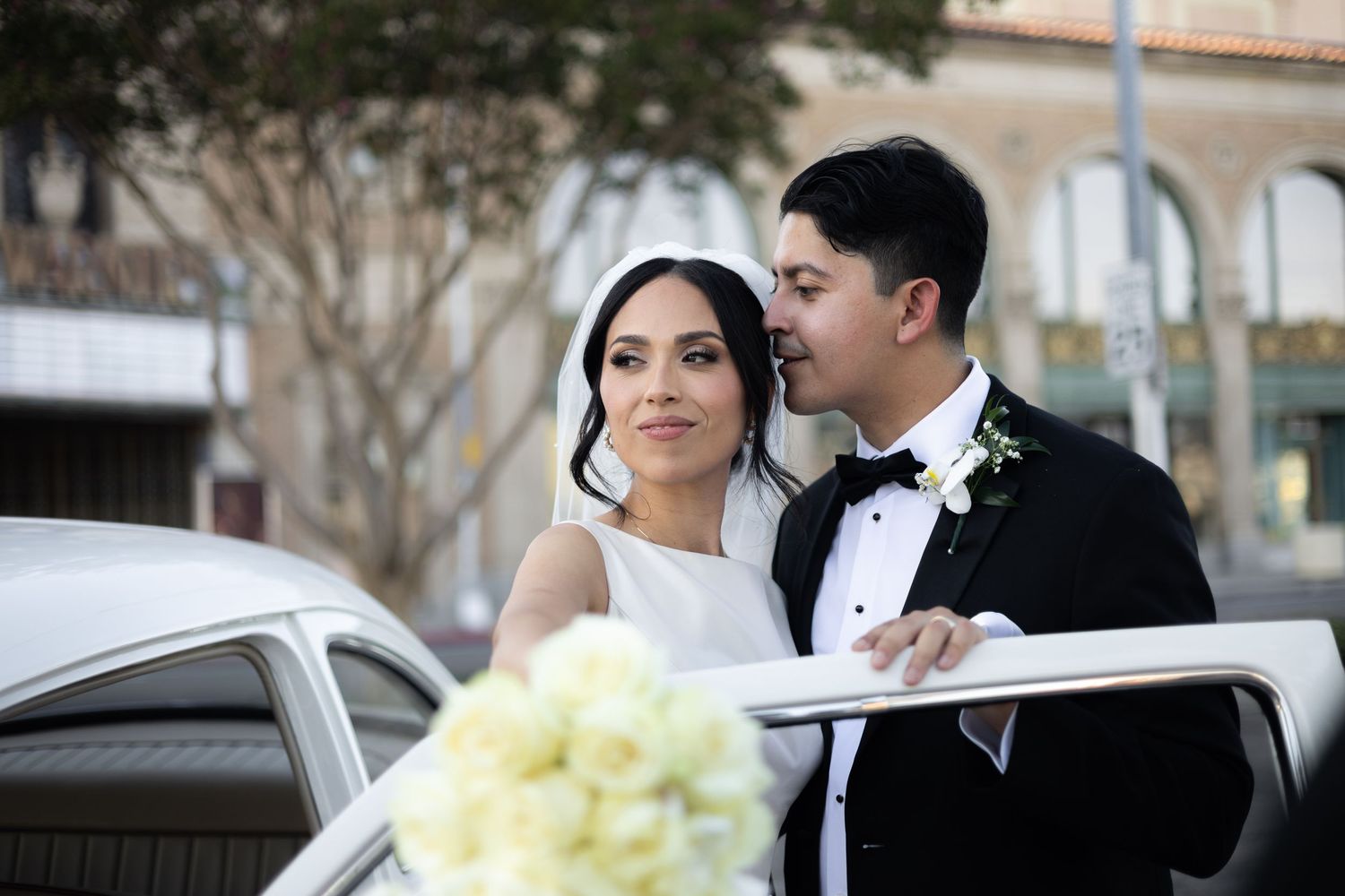 Elegant wedding couple poses with classic white vintage car under city trees.