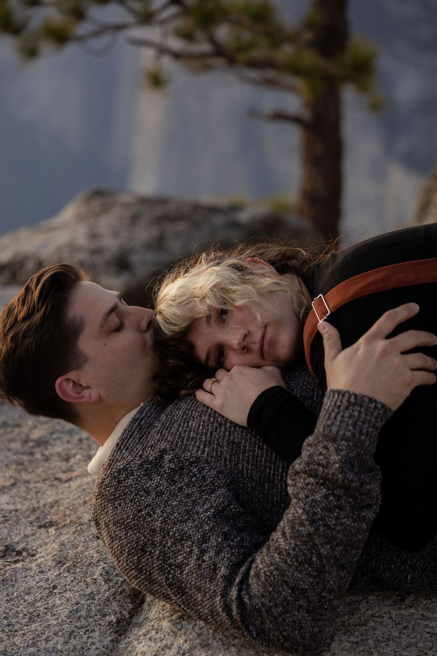 Two people rest intimately on rocky ground with mountain scenery behind them.