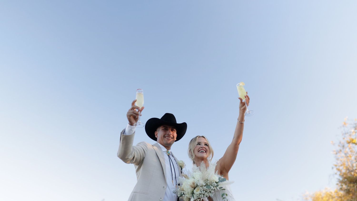 Bride and groom toasting with champagne glasses outdoors under a clear blue sky at their wedding celebration.