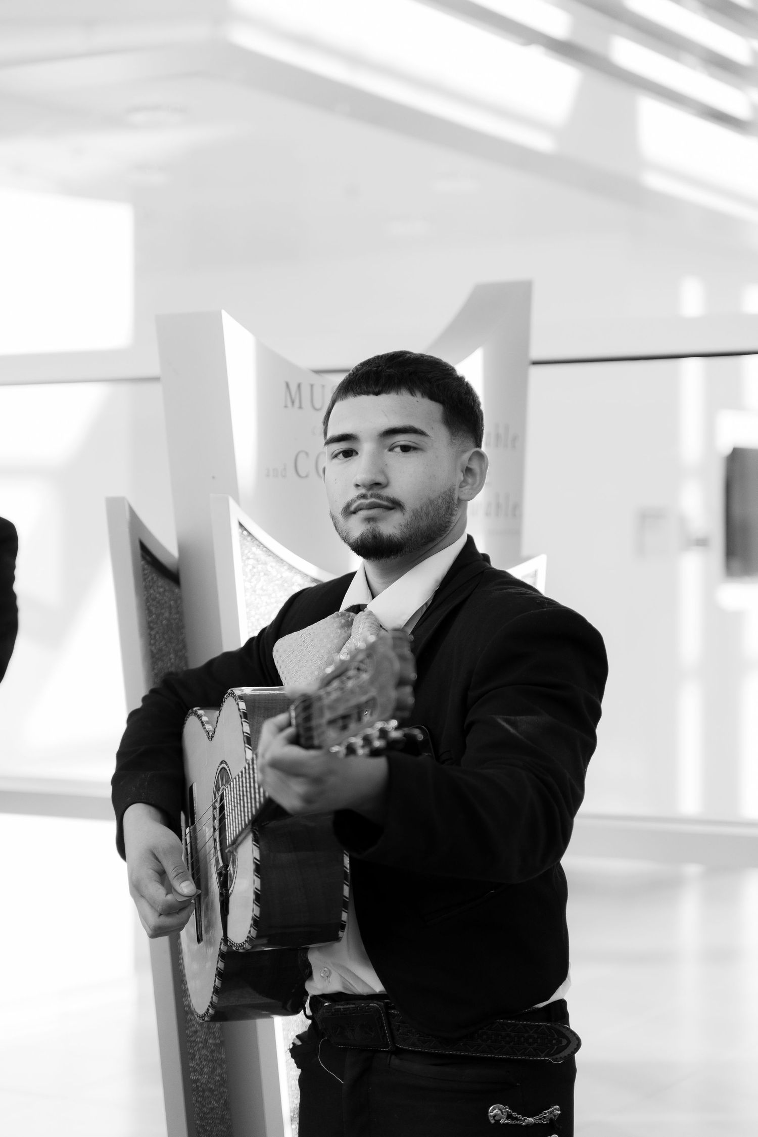 Musician in formal attire plays acoustic guitar in bright white room.