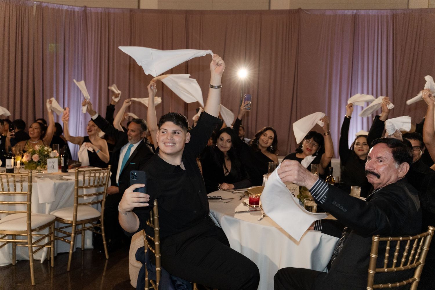 Wedding reception guests celebrating with raised arms and napkins in a decorated ballroom.