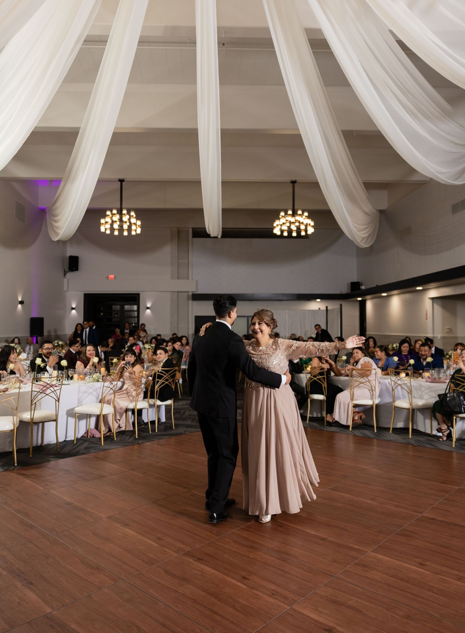 Wedding reception dance floor with guests seated at tables and white draped ceiling decor.