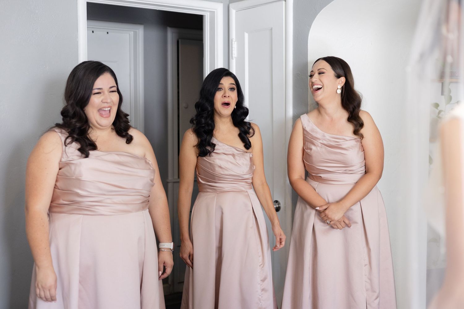 Bridesmaids in matching blush dresses share genuine laughter during wedding preparations.