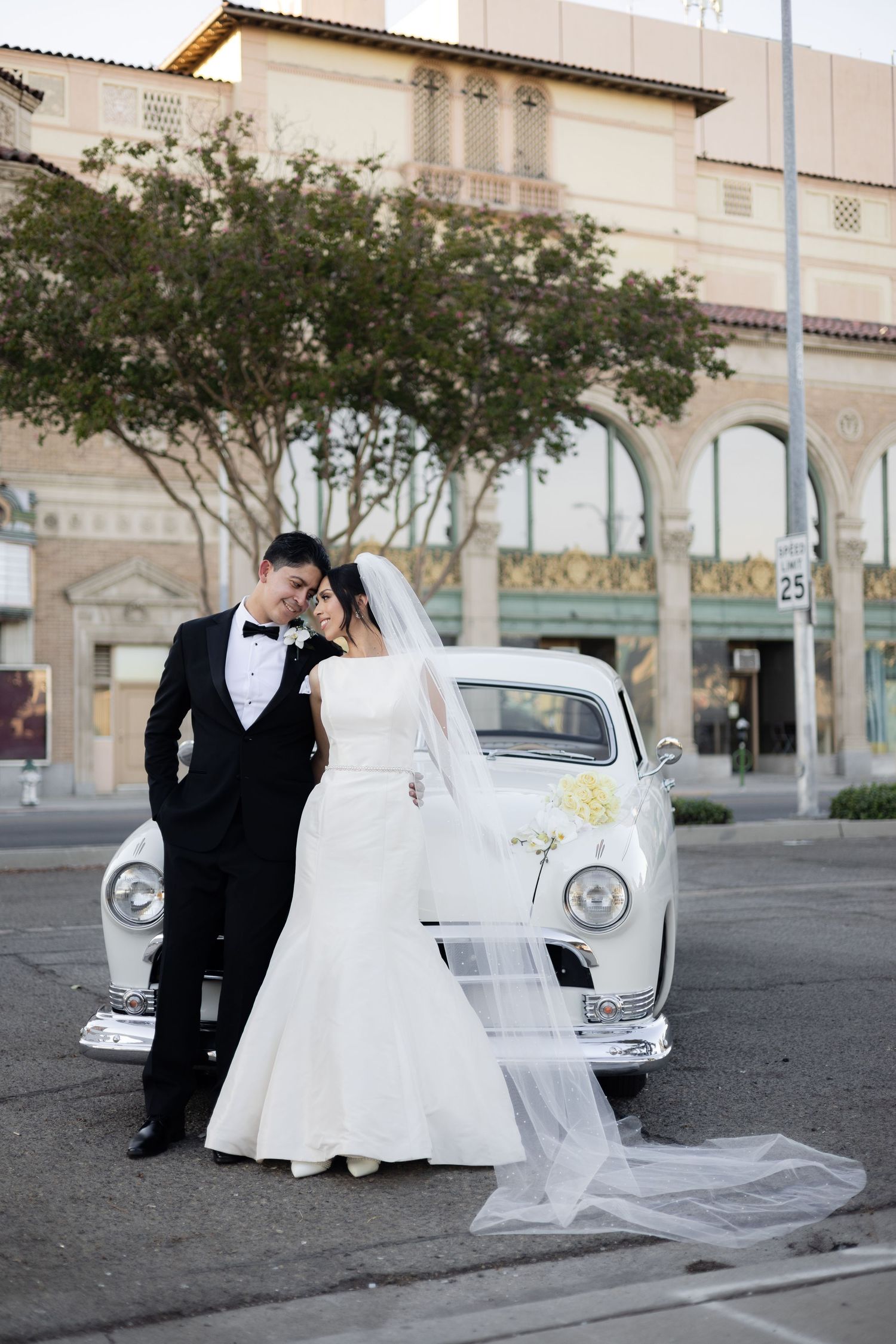 Wedding couple poses with classic white car in front of historic building.