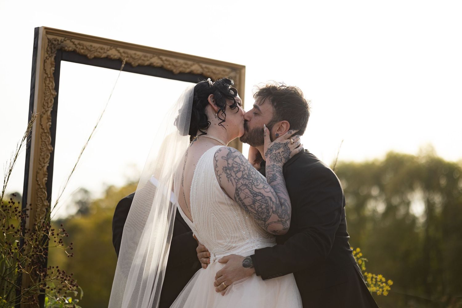 Couple shares intimate kiss under wooden arch during golden hour wedding ceremony.