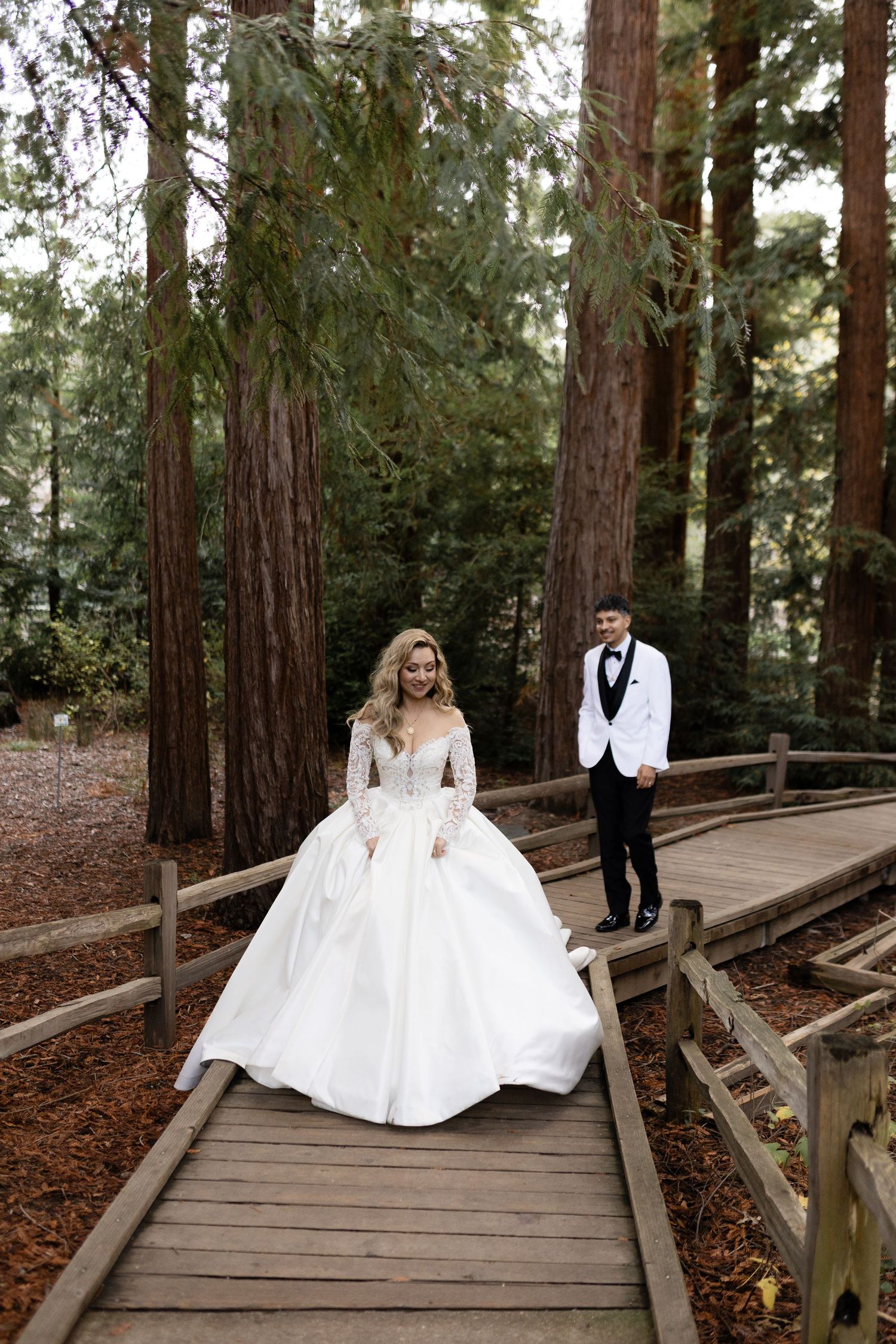 A wedding couple poses separately on different levels of a wooden walkway in a forest setting.