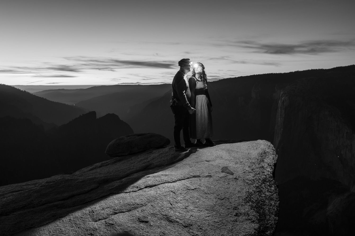 Black and white photo of couple standing on mountain cliff during sunset.