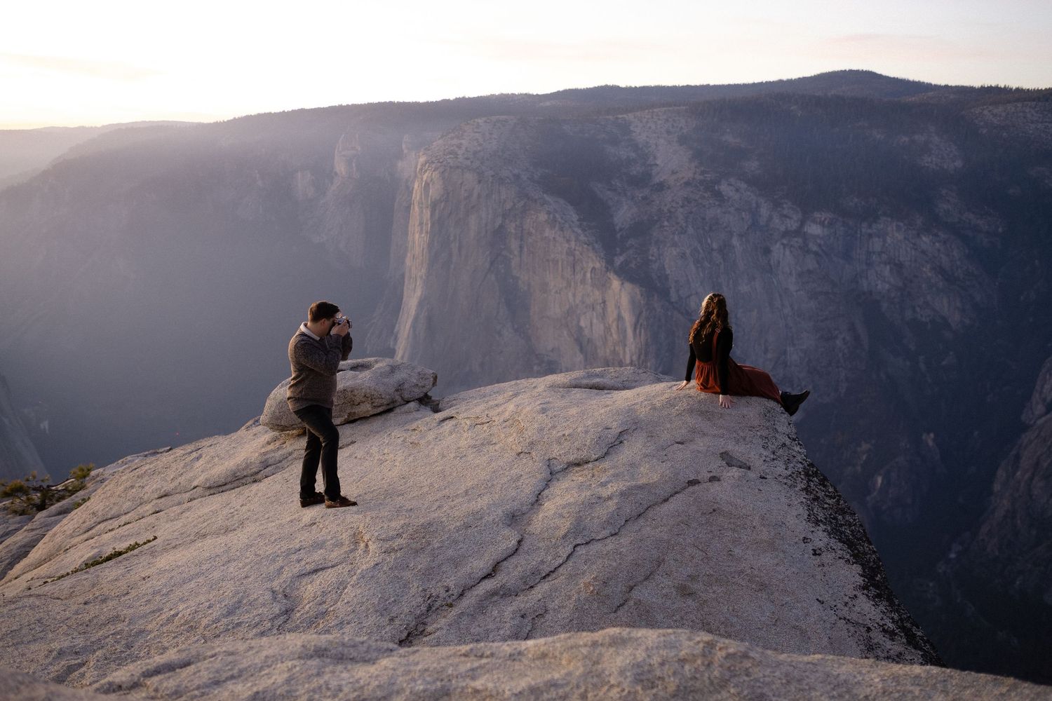 Two people relaxing on edge of cliff with panoramic mountain views in background.