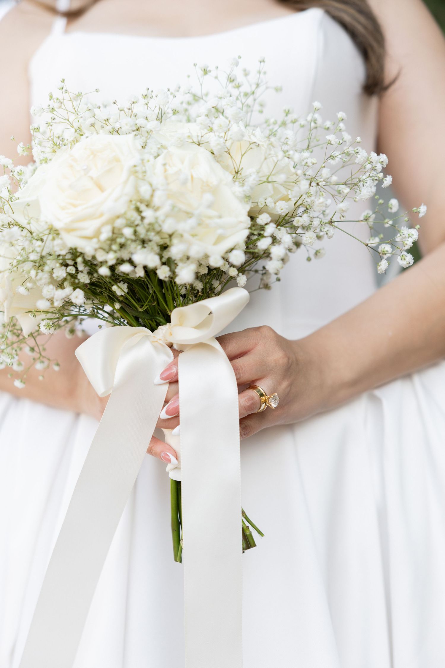 Bride in white dress holding elegant wedding bouquet of white roses and baby's breath tied with satin ribbon bow.
