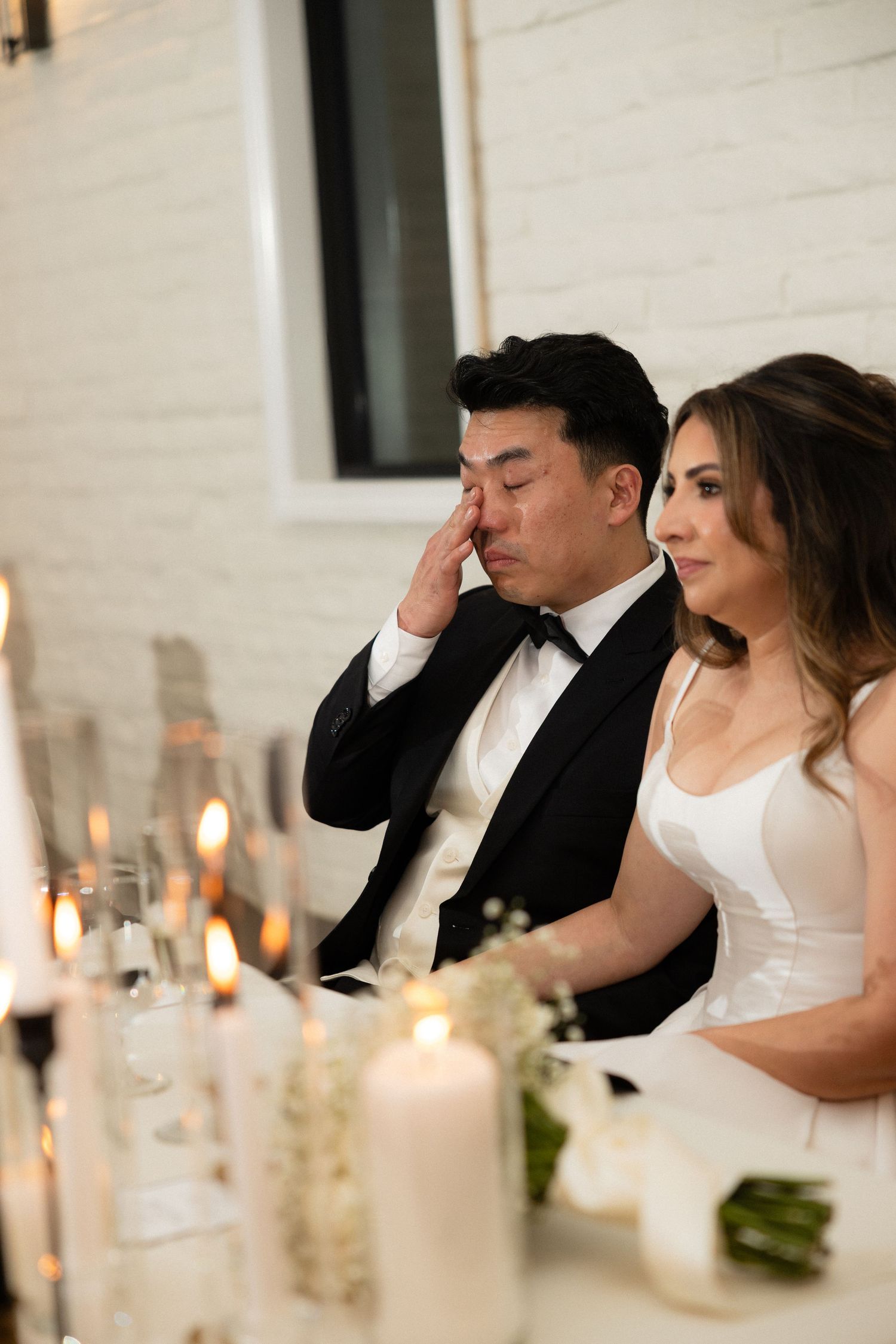 Groom wiping tears at wedding reception table with candles while bride sits beside him.