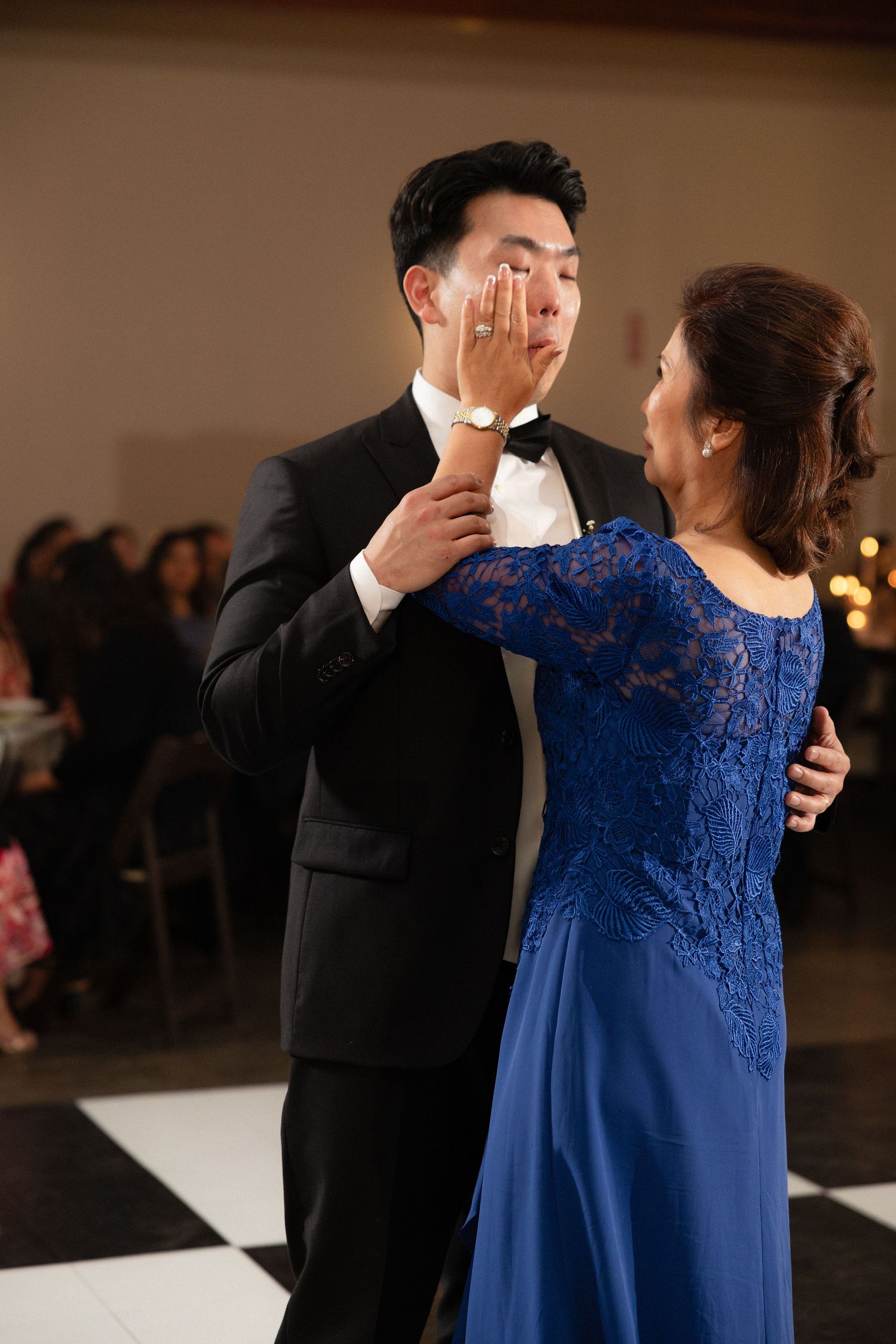 Groom in black tuxedo wipes tears during emotional mother-son dance, mother wearing blue lace gown.