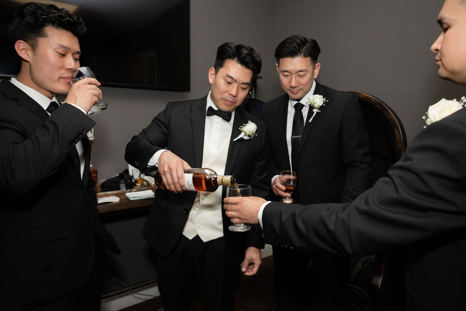 Groom in tuxedo pouring whisky for groomsmen during pre-wedding celebration in a wedding venue.