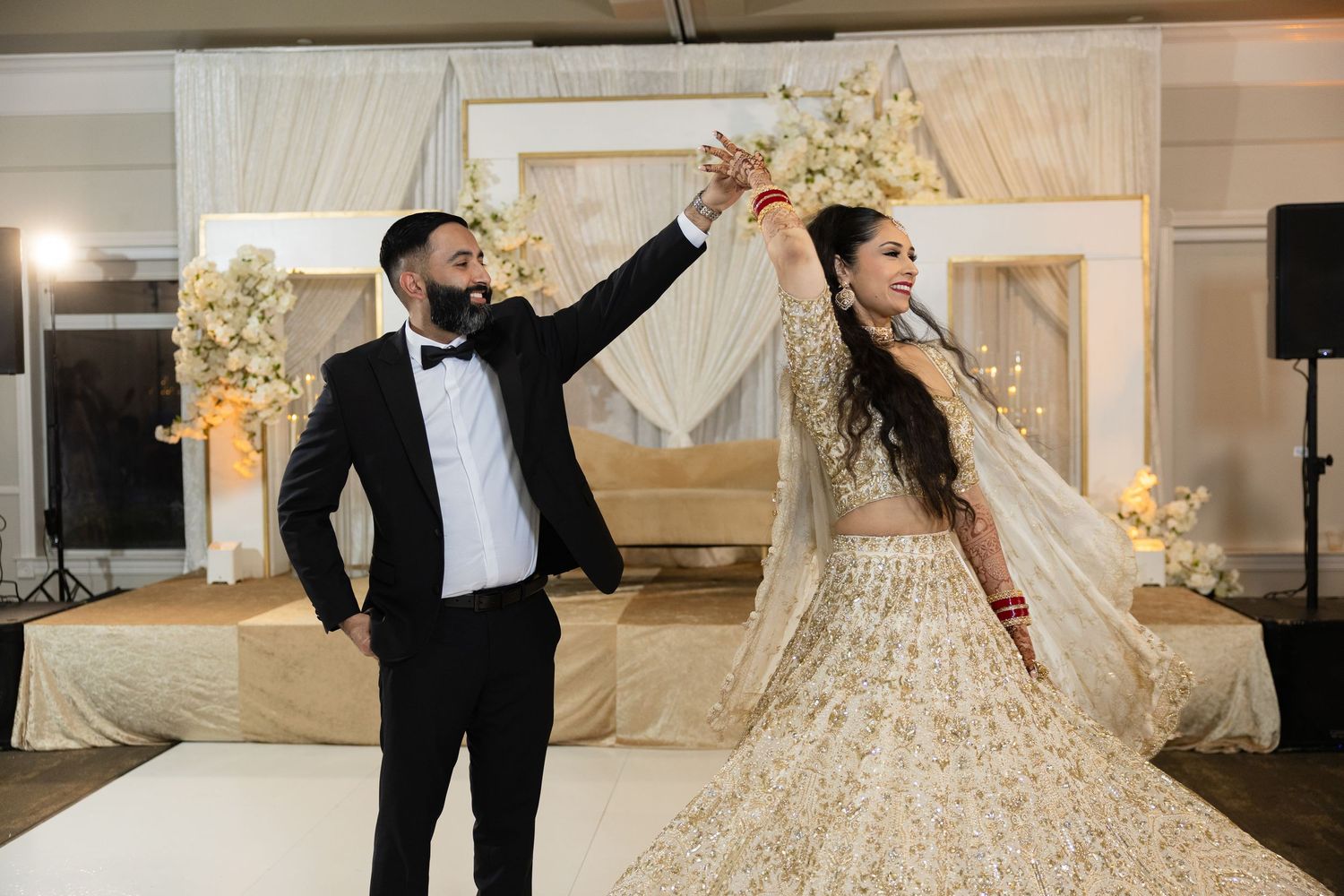 A couple performs a celebratory dance move on a white stage with floral decorations at a formal event.