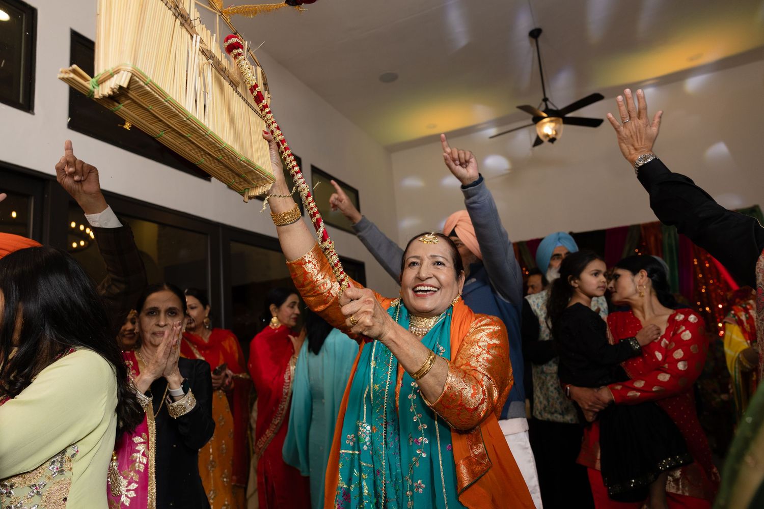 Wedding guests dance with raised hands during a lively celebration in an indoor venue with ceiling fans.