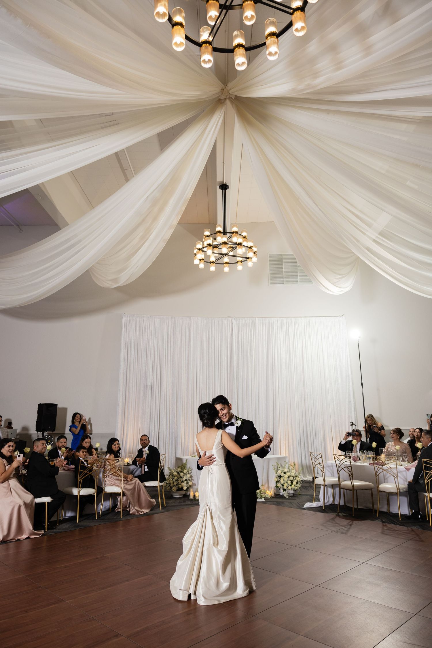 Wedding couple dances beneath elegant white fabric ceiling draping and chandelier lighting.