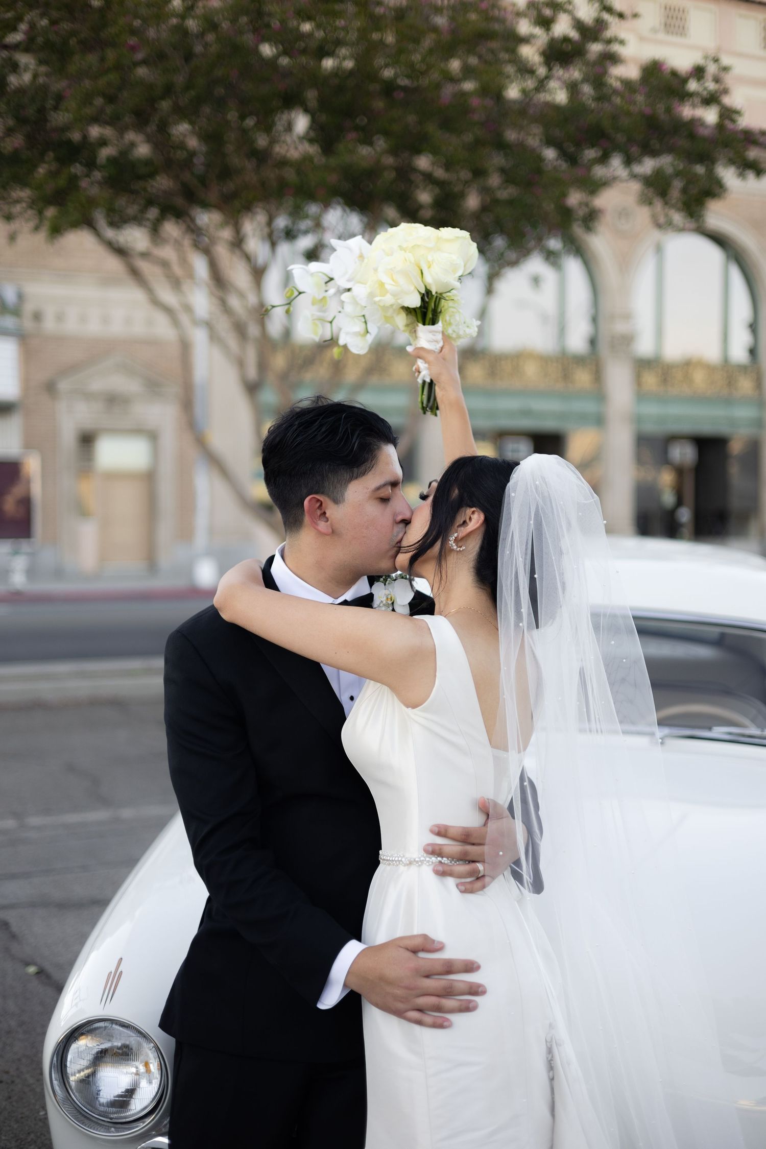 Couple in wedding attire embracing by classic white car.