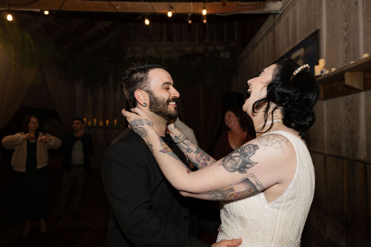 Couples dance and celebrate at a rustic barn wedding reception under warm string lighting.