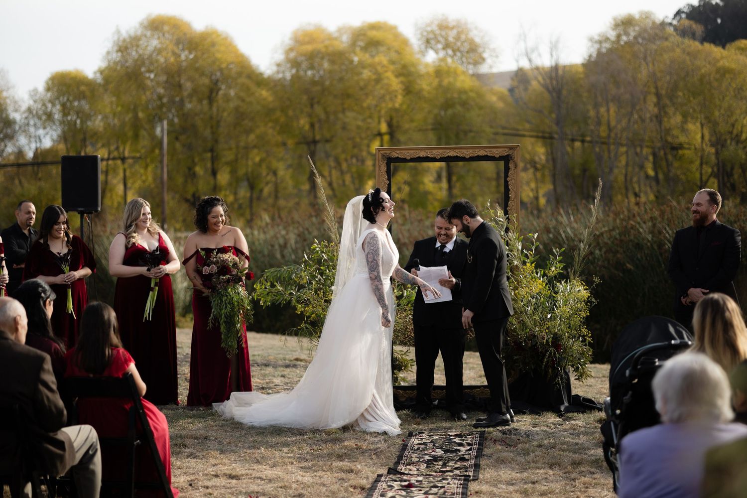 An outdoor wedding ceremony takes place under a rustic wooden arch with autumn trees in the background.