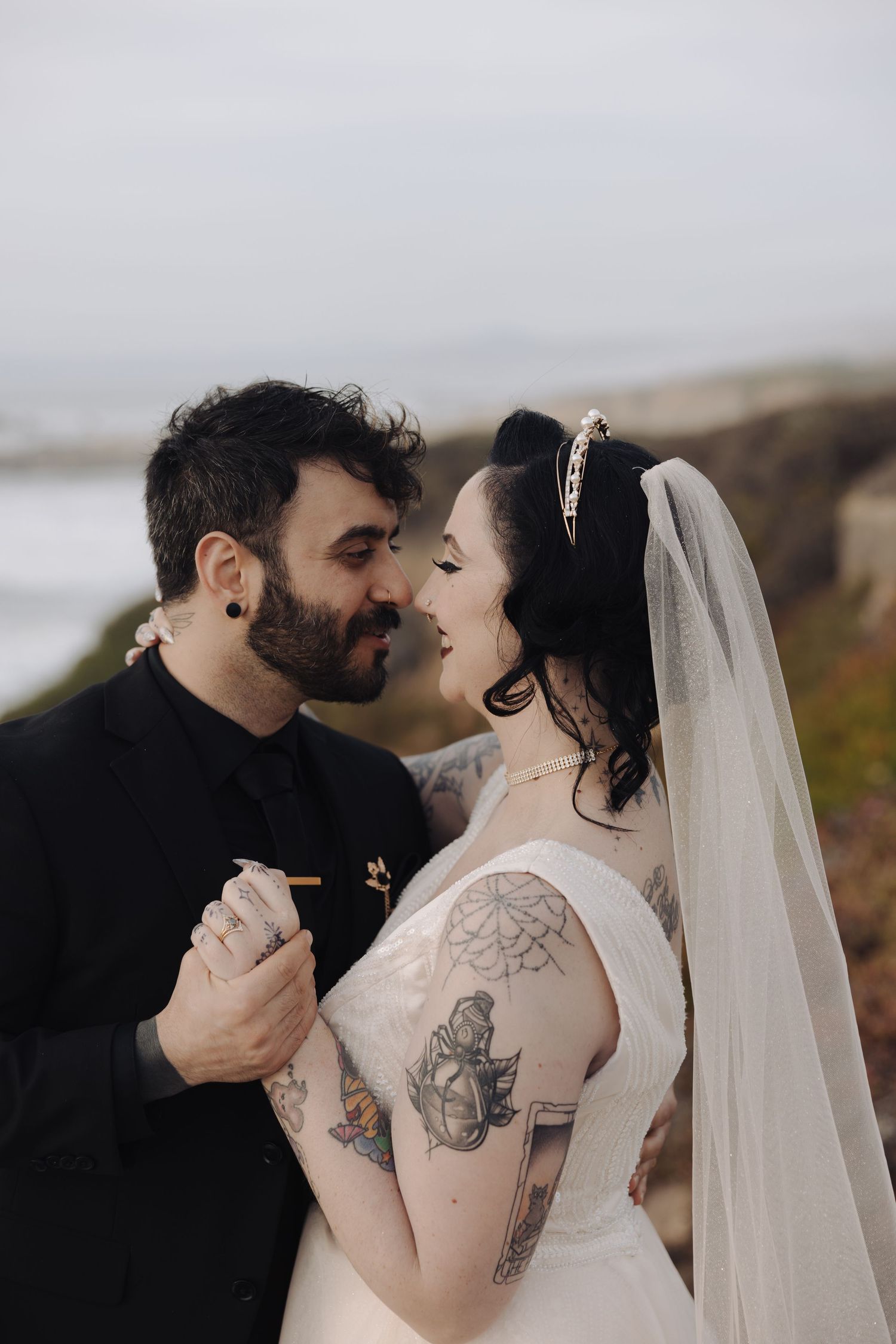Newlyweds in black and white attire share a tender moment against a misty coastal landscape.