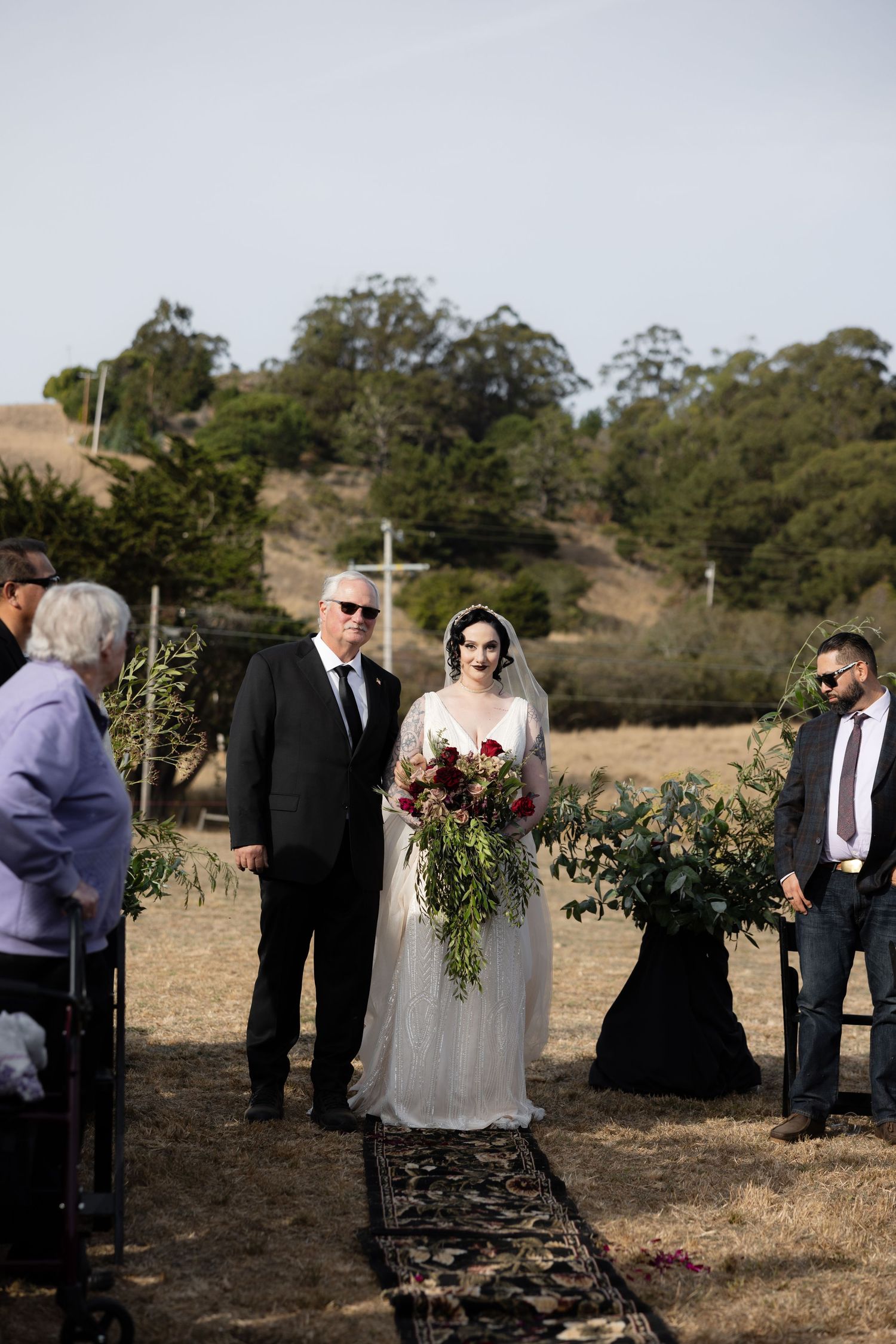 Wedding ceremony takes place outdoors with guests gathered around a wooden arch in a rustic field setting.