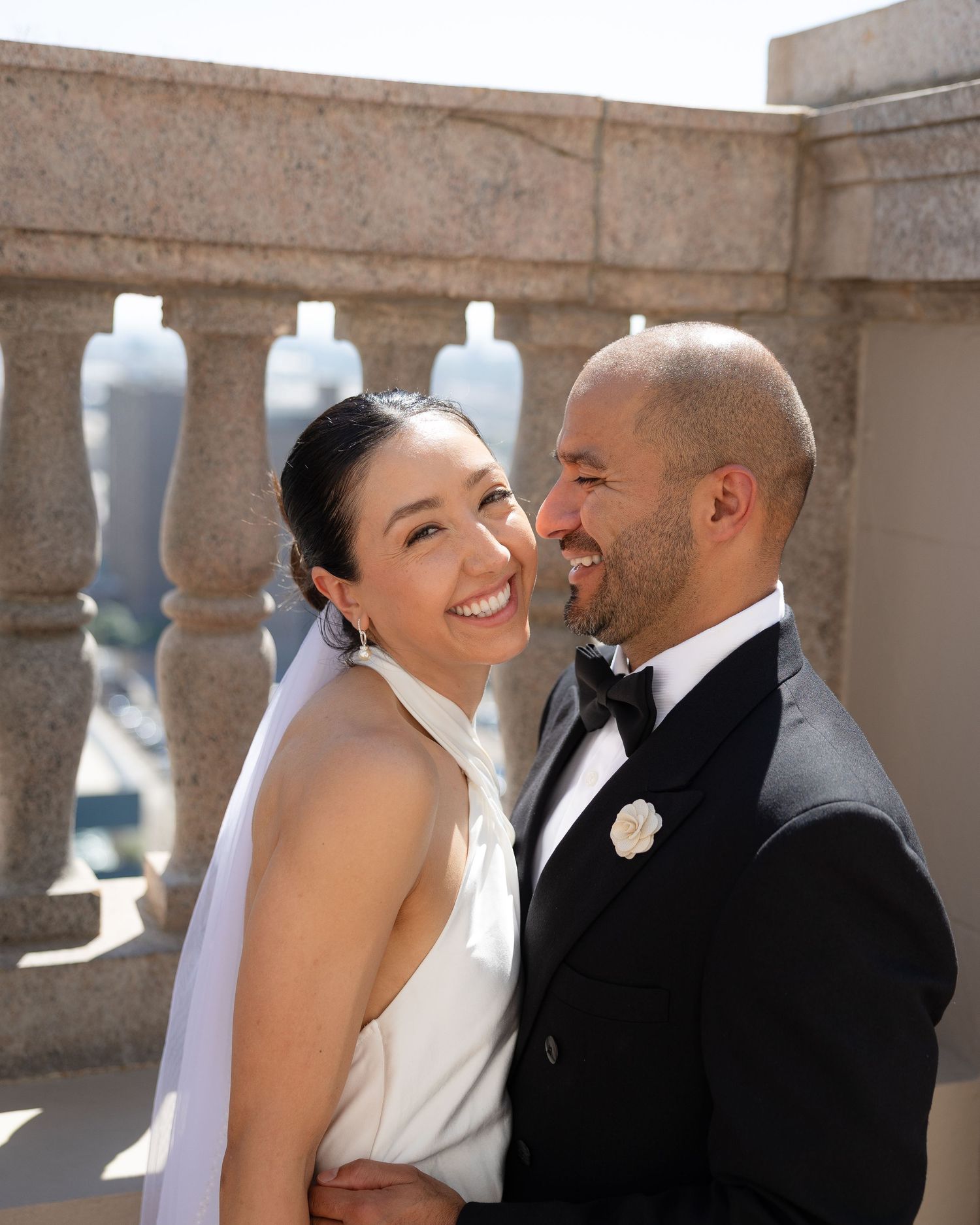 A smiling bride in a white halter gown and veil poses with her groom in a black tuxedo on an elegant stone terrace.
