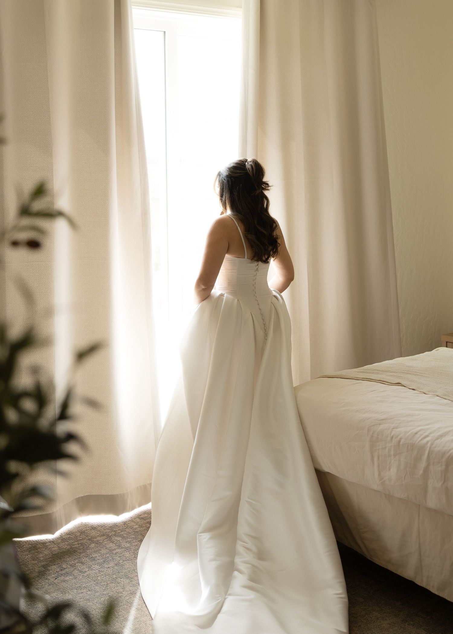Bride in strapless white ball gown standing at sunlit window with flowing skirt and veil.