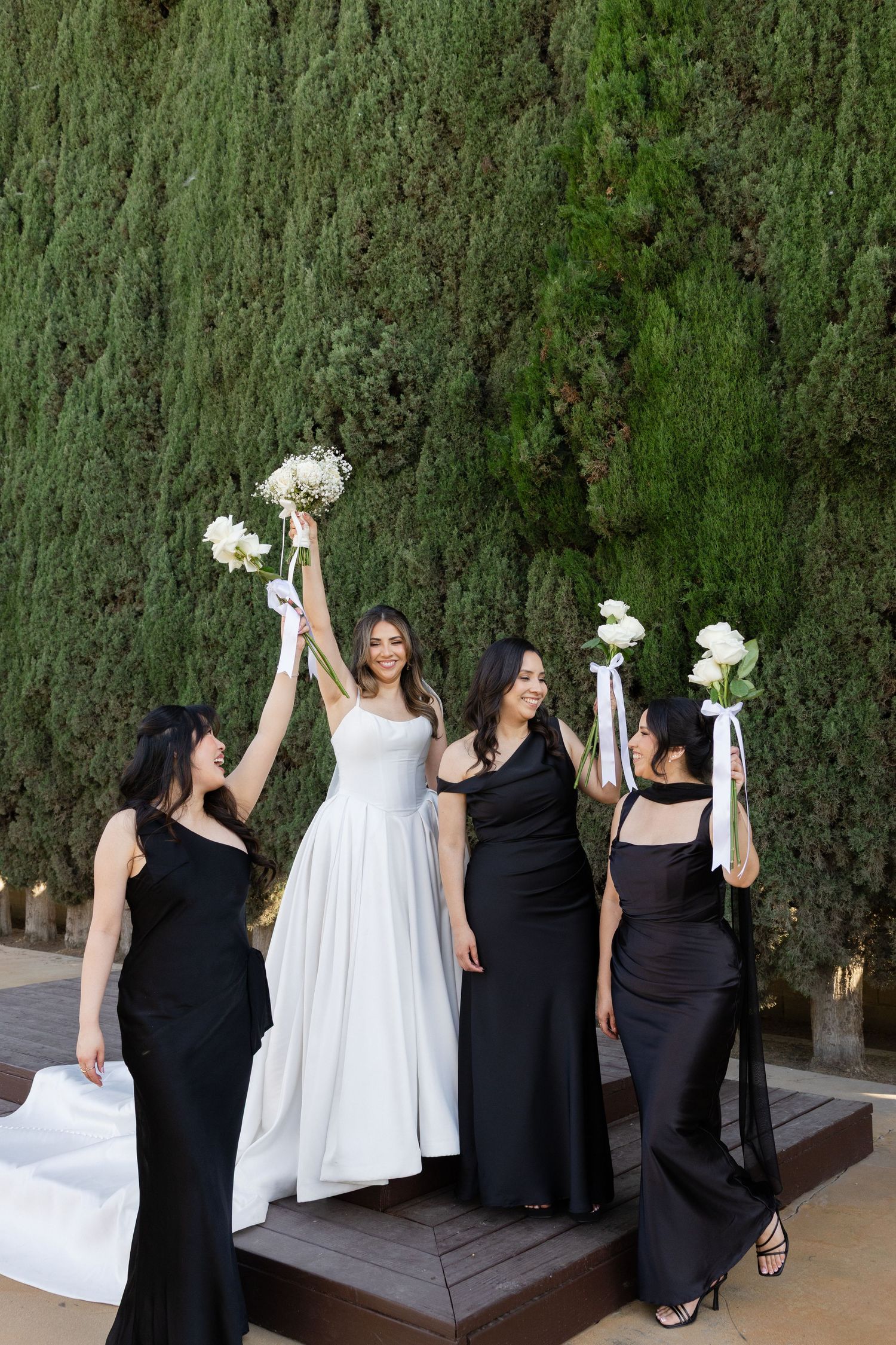 Bride in white gown with three bridesmaids in black dresses raising bouquets outdoors near green cypress trees.