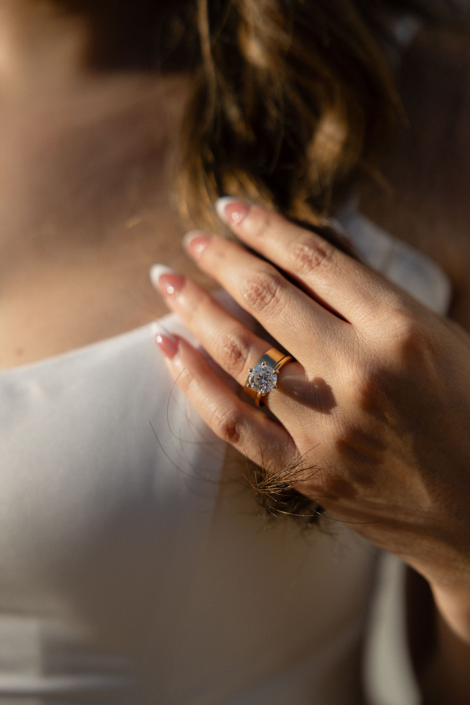 Close-up of a woman's hand wearing a gold diamond engagement ring, resting on her shoulder in warm sunlight.