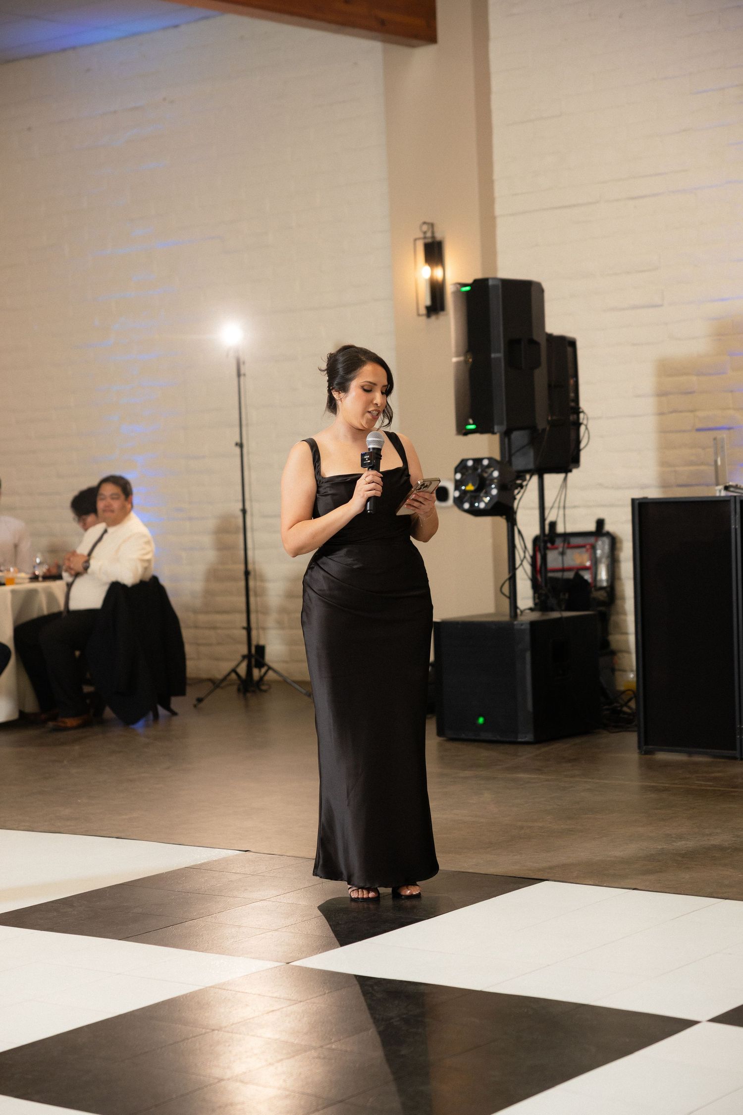 Woman in black gown giving a speech with microphone on checkered dance floor at elegant wedding reception.