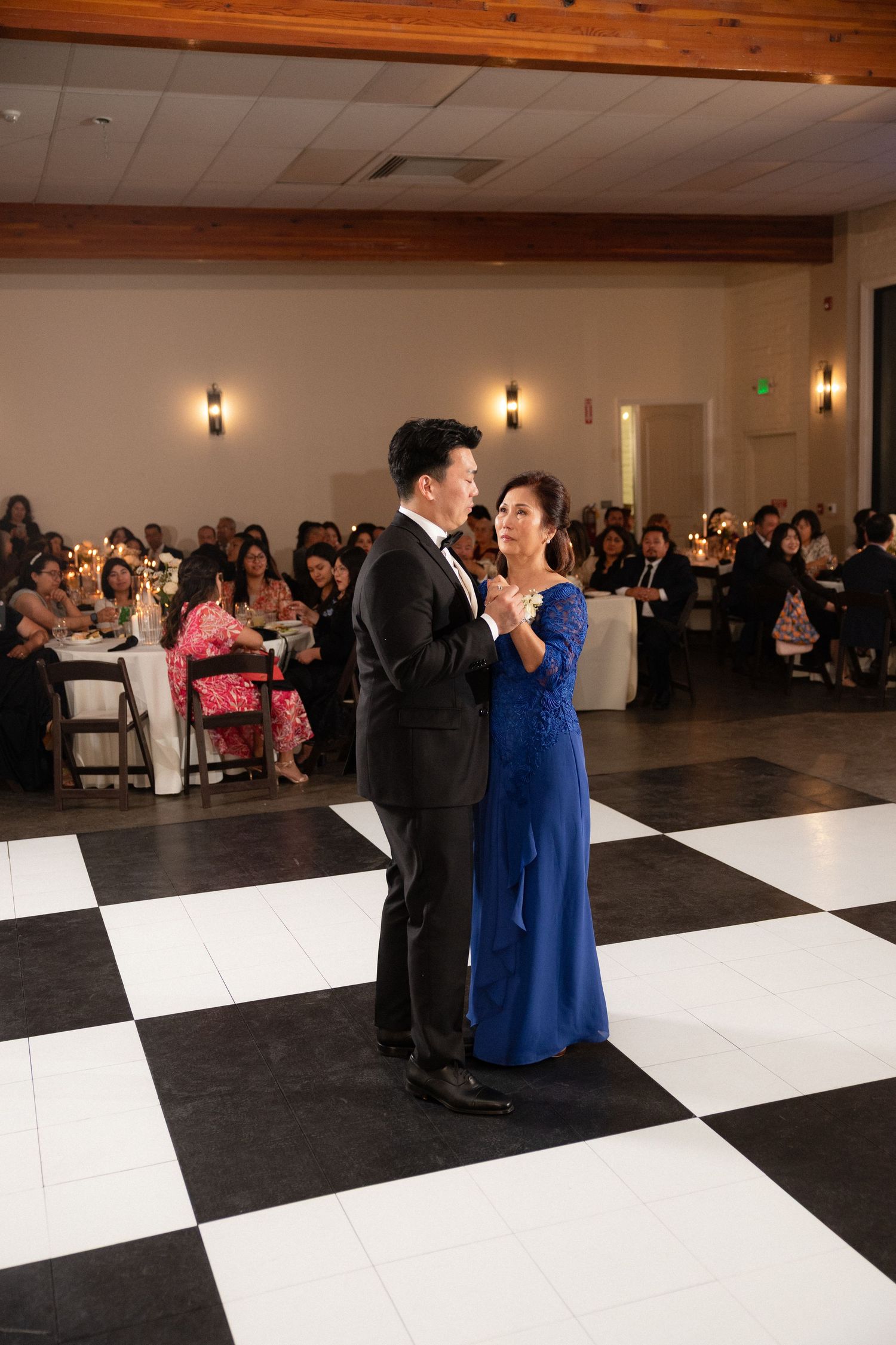 Groom dancing with mother in blue gown on checkered dance floor at wedding reception.