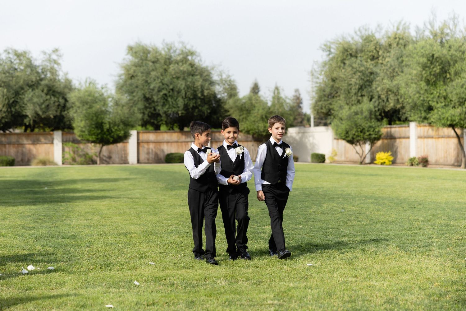 Three boys in matching black suits and vests walk together on a green lawn at an outdoor event.