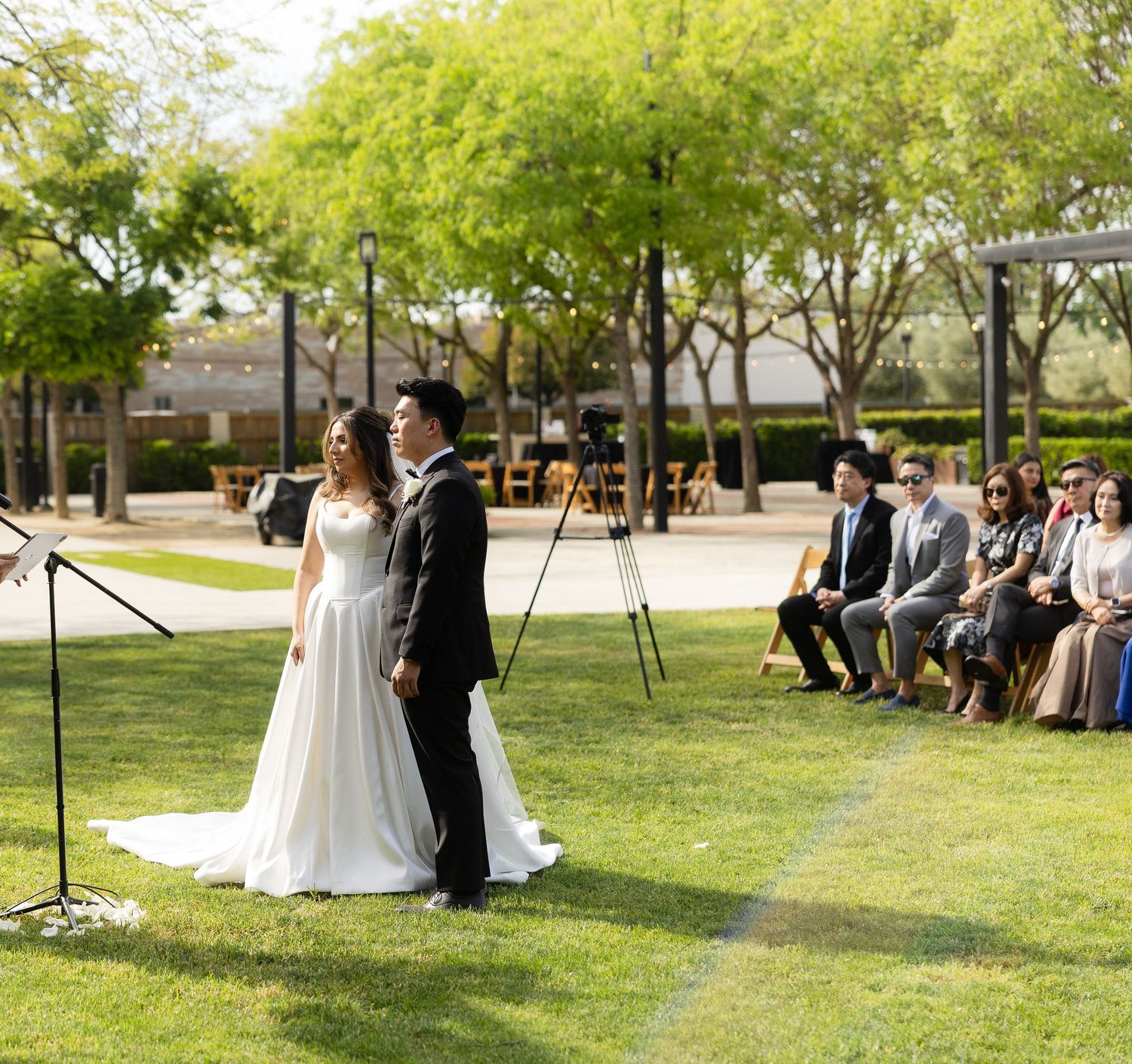 Couple exchanges vows at outdoor wedding ceremony on green lawn surrounded by seated guests and lush trees.