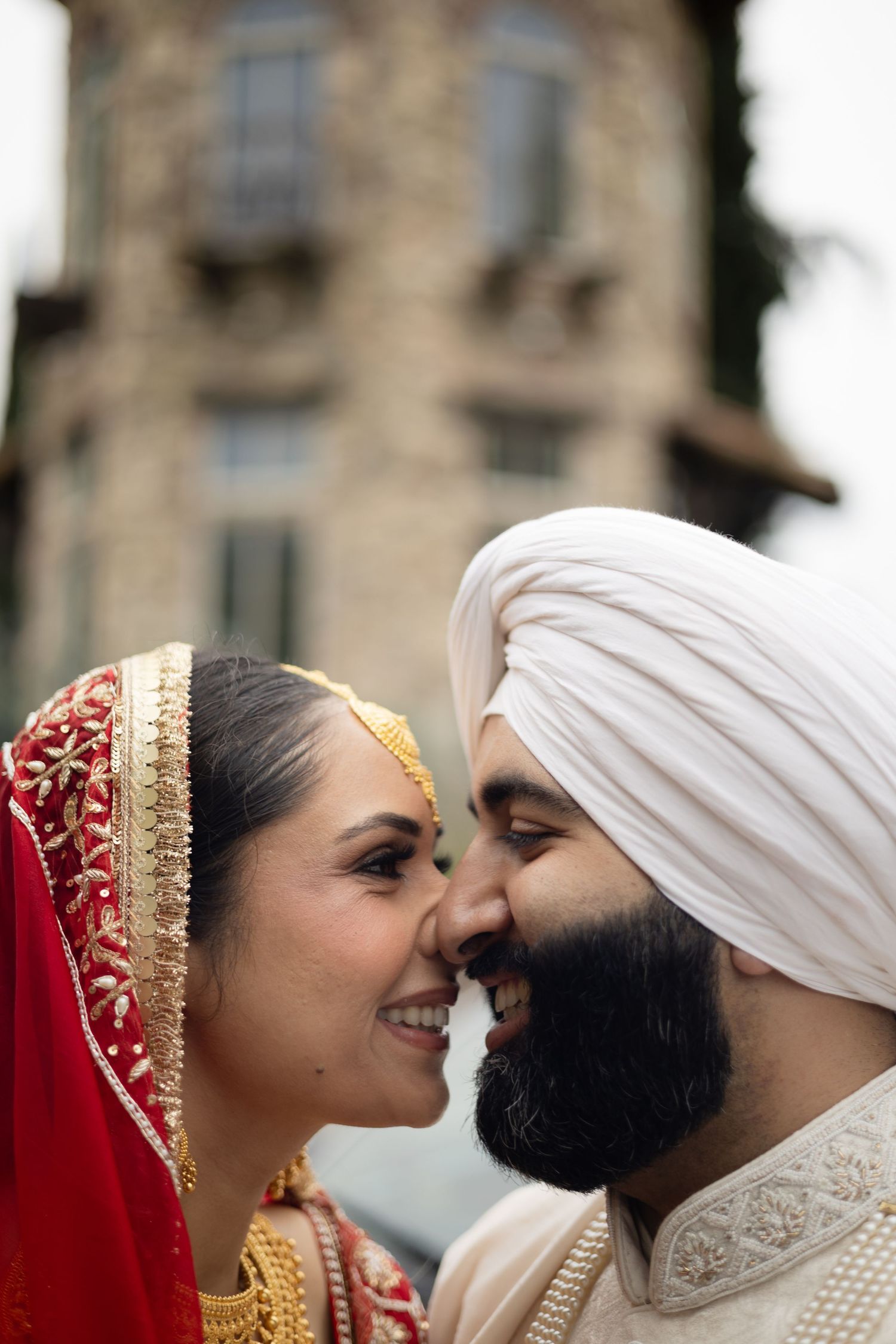 A romantic moment between a couple in traditional Sikh wedding attire against a stone building backdrop.