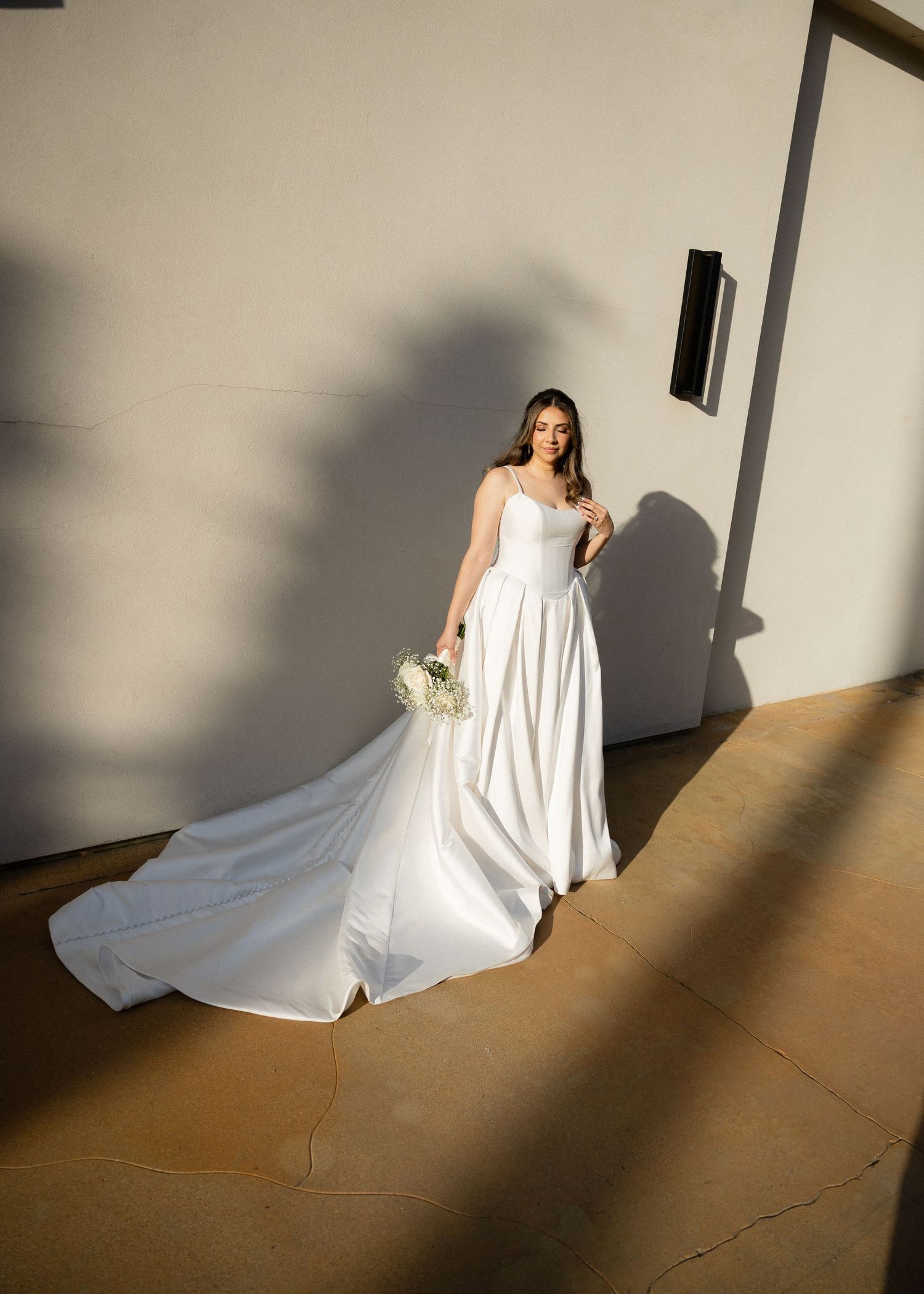 Bride in elegant white satin ball gown with long train holding bouquet, standing in dramatic sunlight against a neutral wall.