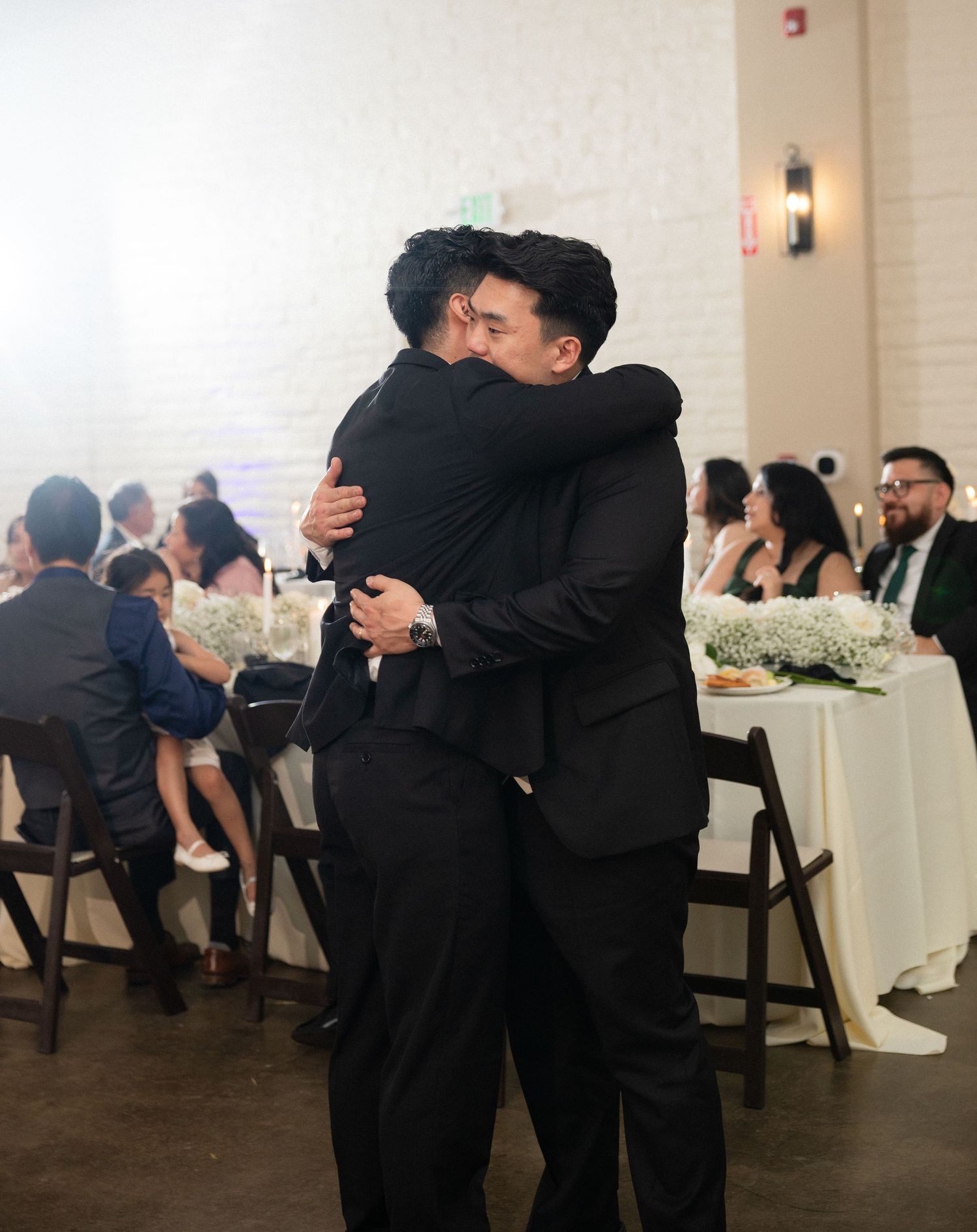 Two men in black suits share an emotional hug at a wedding reception with guests seated in background.