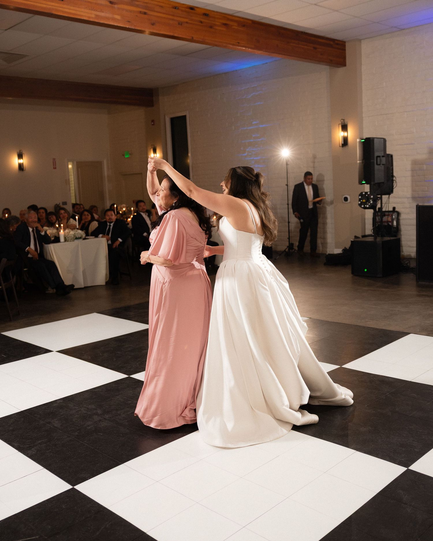 Bride dancing with woman in pink gown on checkered dance floor at wedding reception.