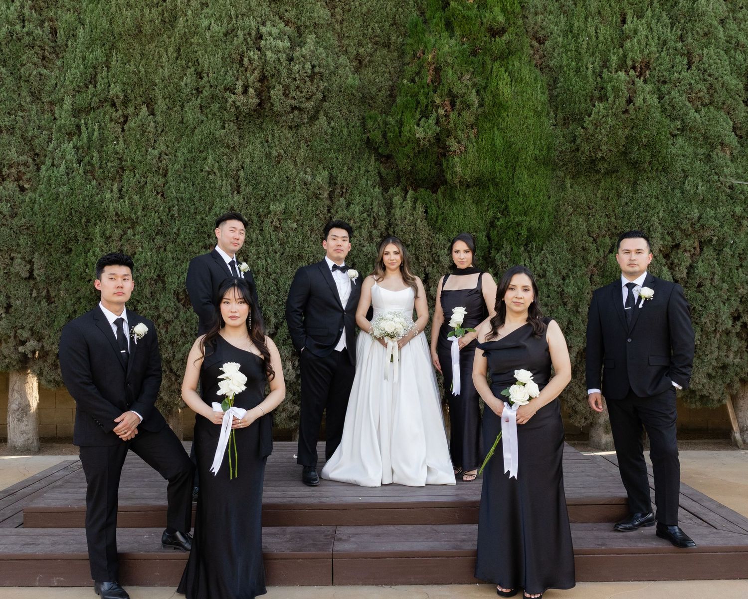 Wedding party in black attire with bride in white gown posing outdoors before a lush green hedge backdrop.