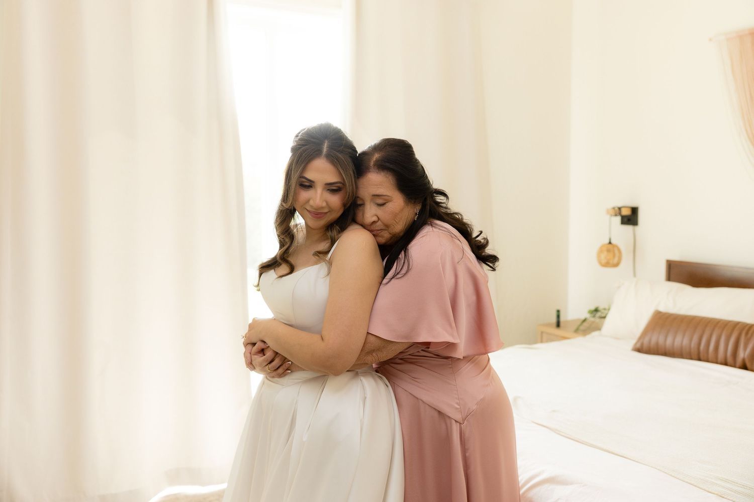 Bride in white gown embraced by woman in pink dress in bright bedroom during wedding preparations.