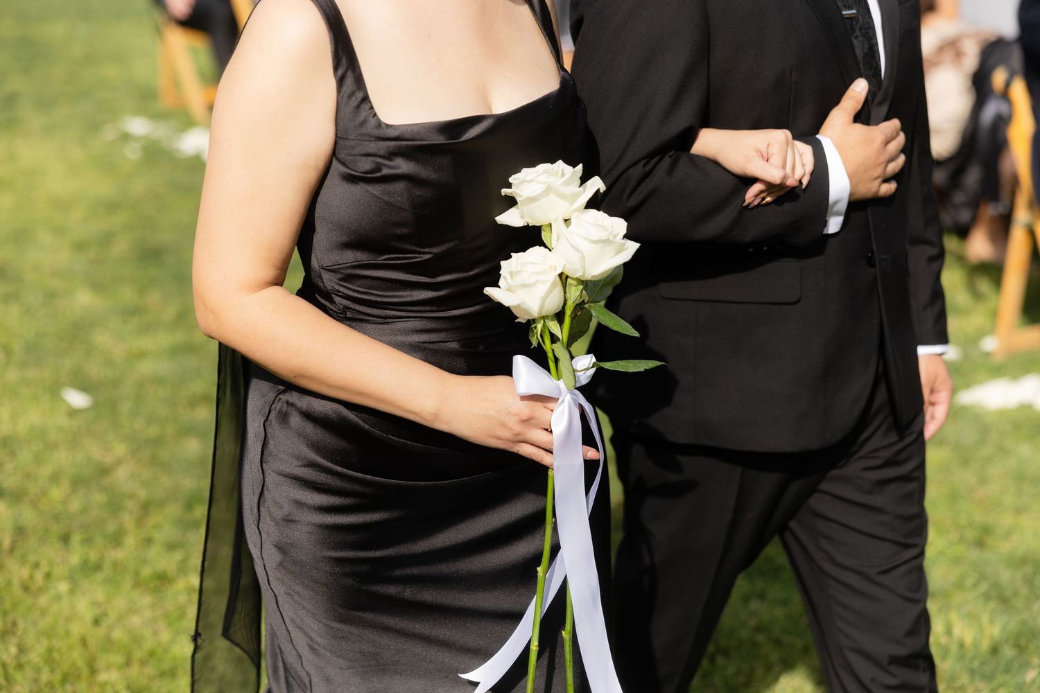 Woman in black dress holding white roses walks with man in black suit at outdoor wedding ceremony.