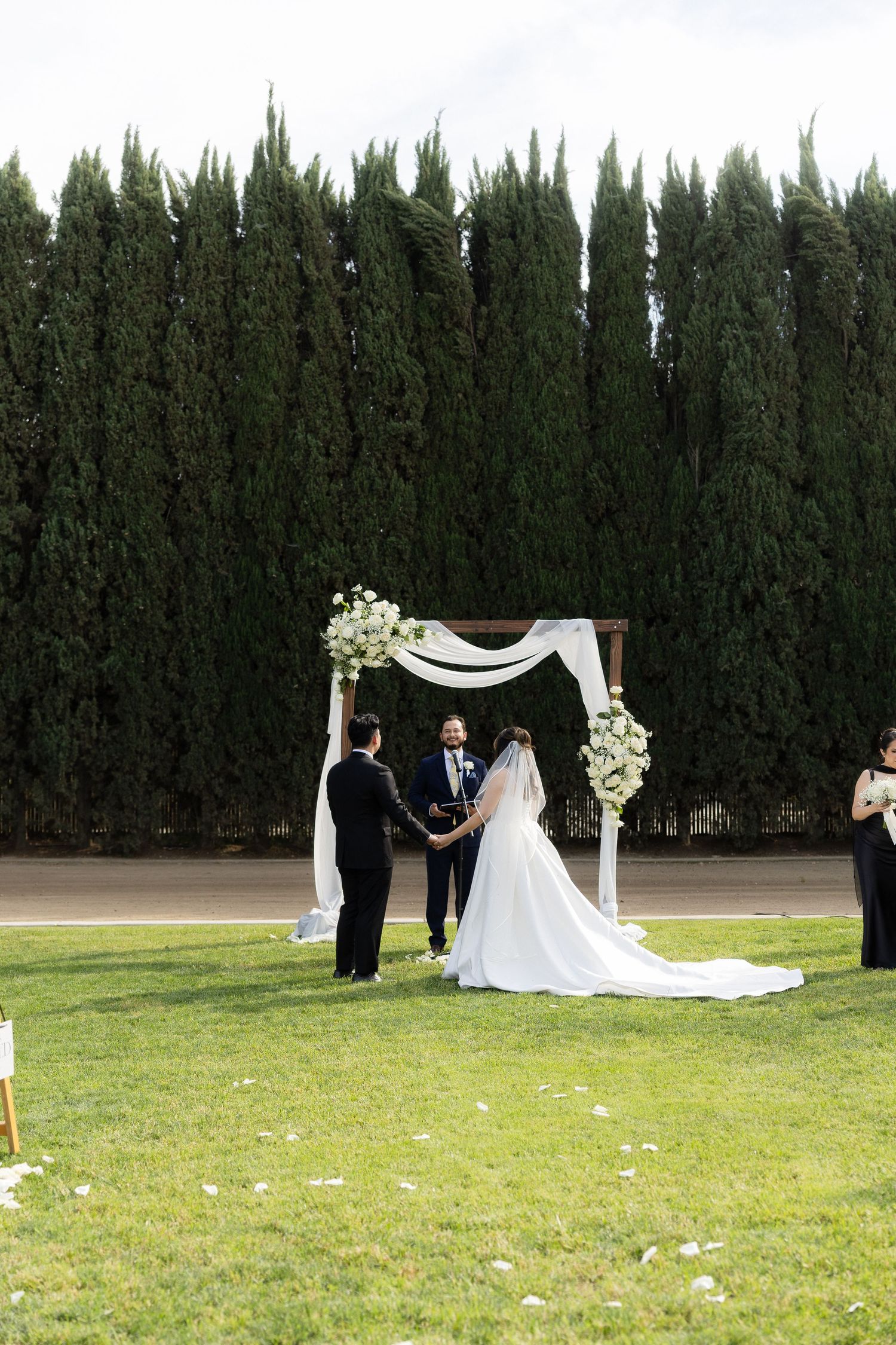 Bride and groom exchange vows at floral wedding arch surrounded by tall cypress trees.