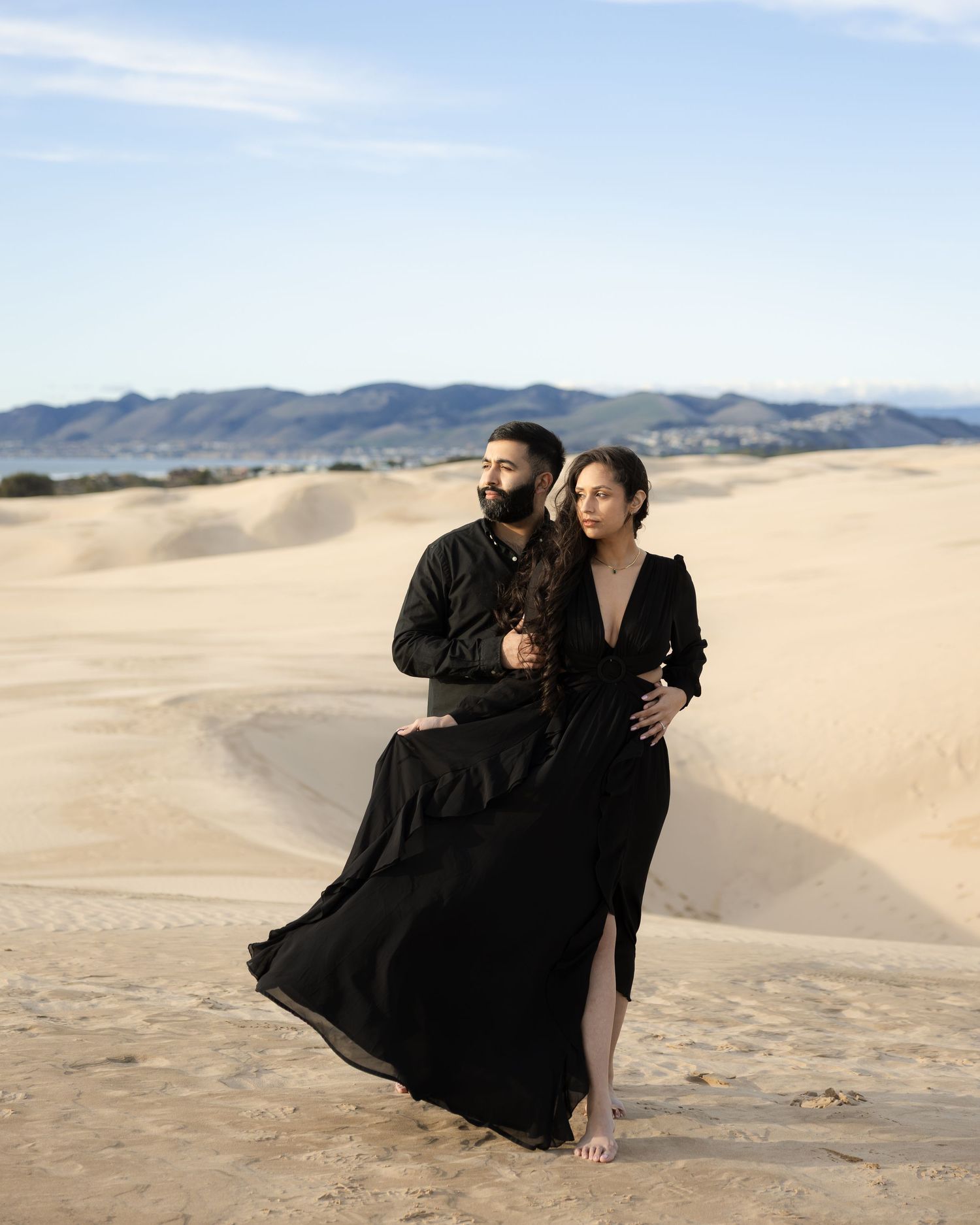 Two people in elegant black outfits pose together against a backdrop of rolling sand dunes and distant mountains.