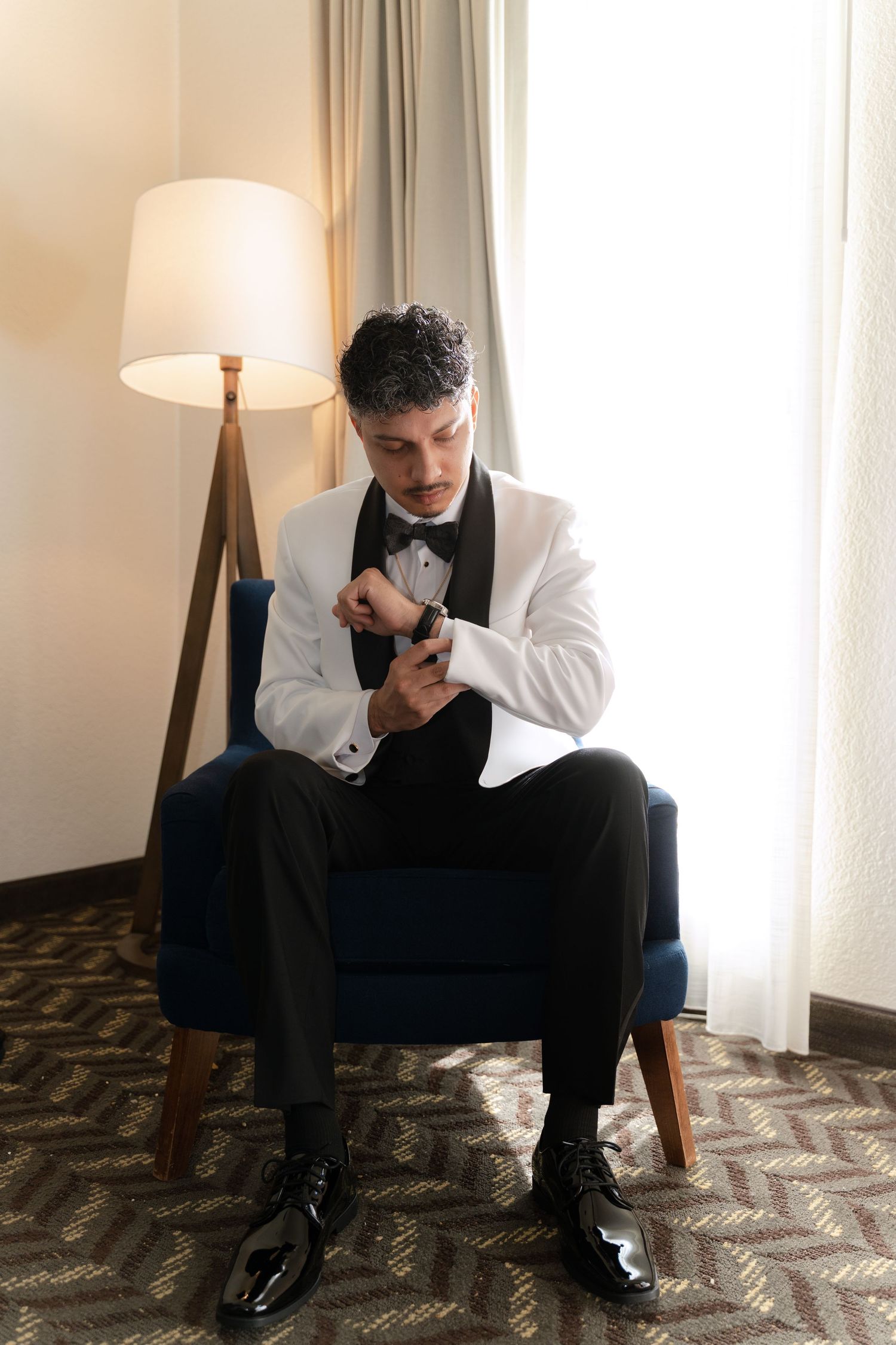 A person in a white tuxedo jacket and black pants adjusts their cuff while sitting in an armchair next to a floor lamp.