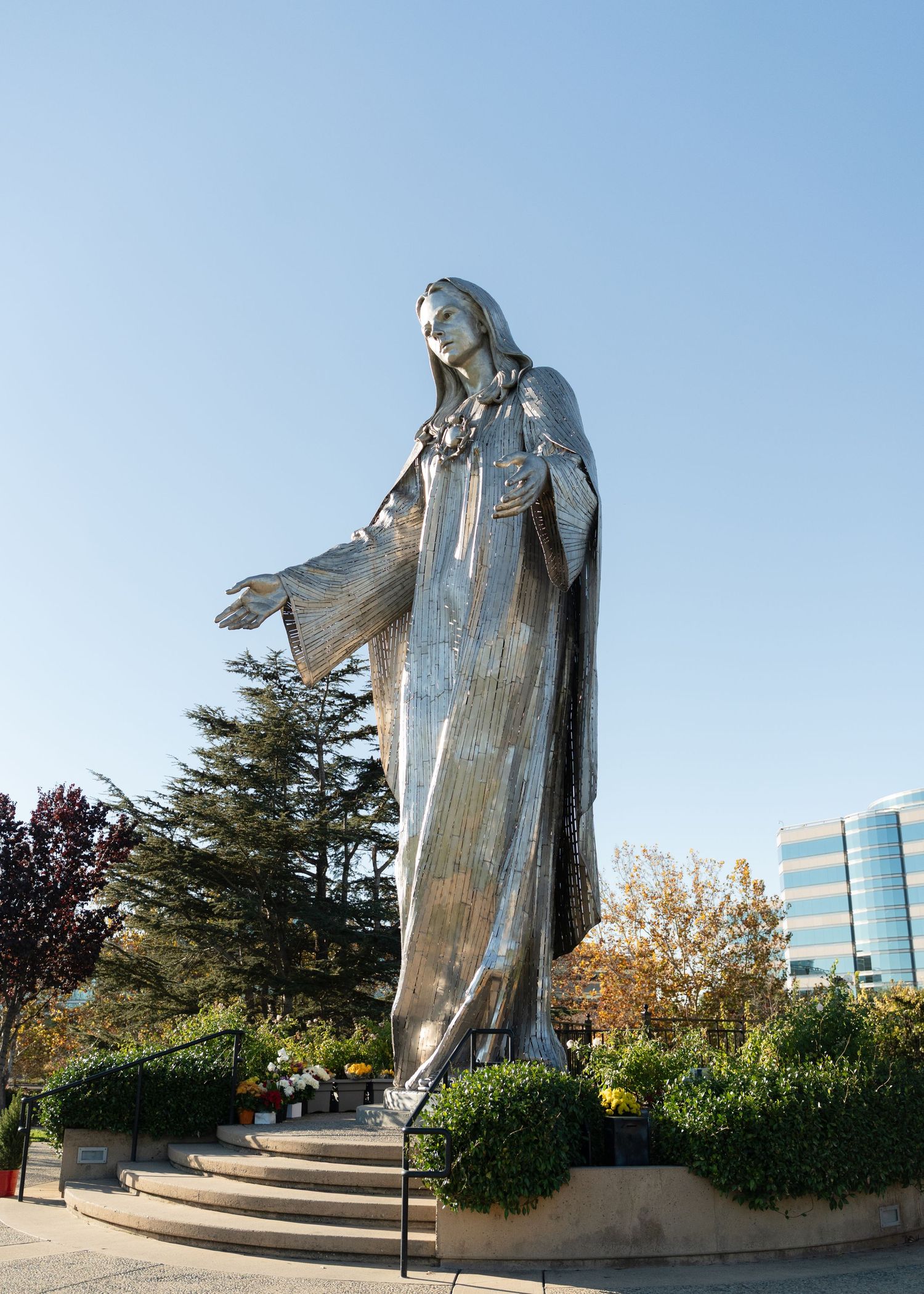 Large silver statue of Jesus with outstretched arms stands against blue sky with trees in background.