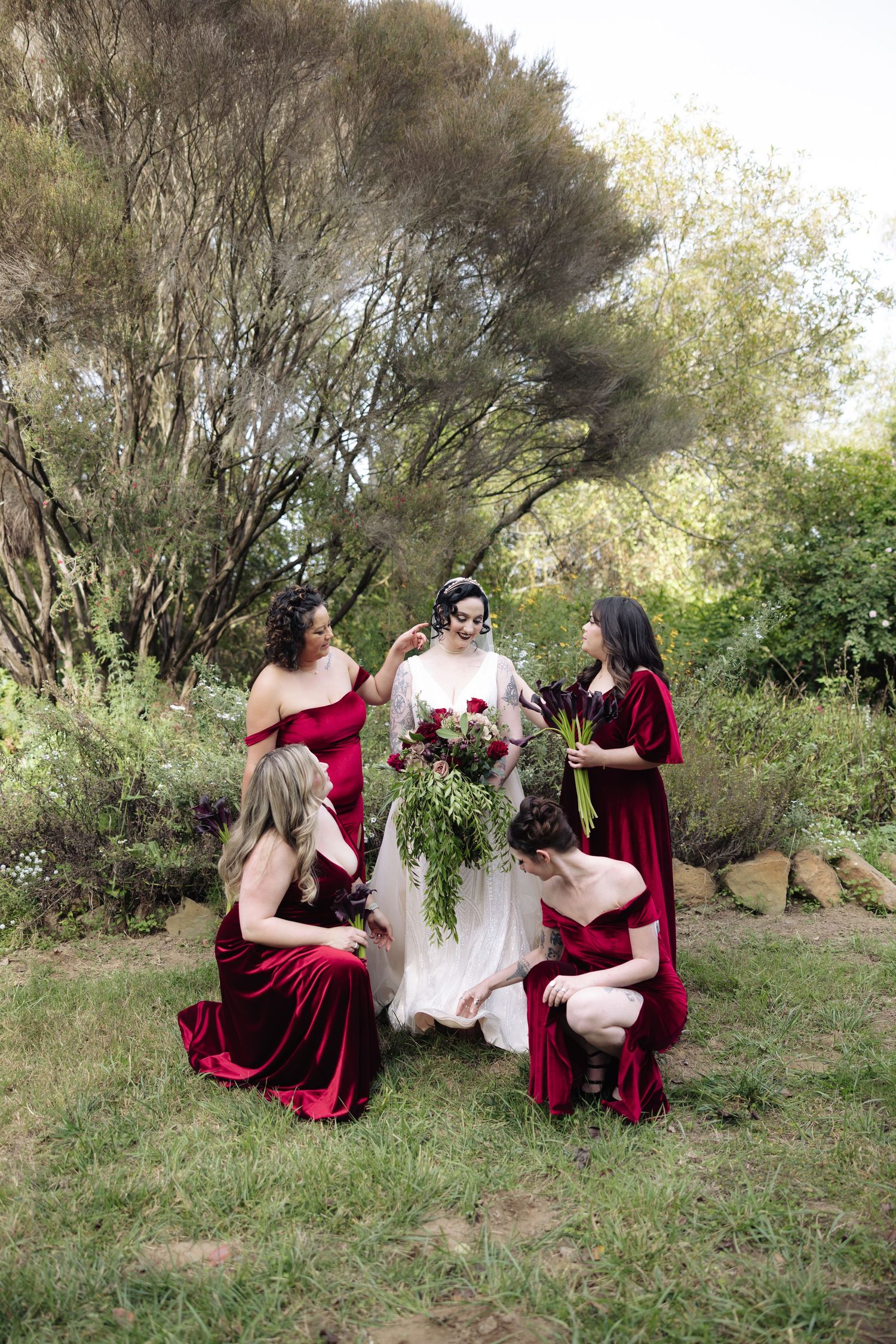 Bridesmaids in burgundy velvet dresses gather around bride in white gown on grassy hillside.