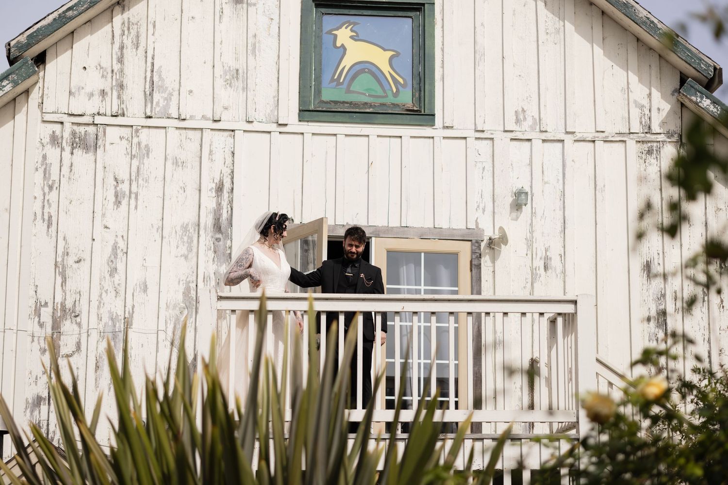 Two people share a moment on a white barn's balcony featuring a folk art deer emblem above.
